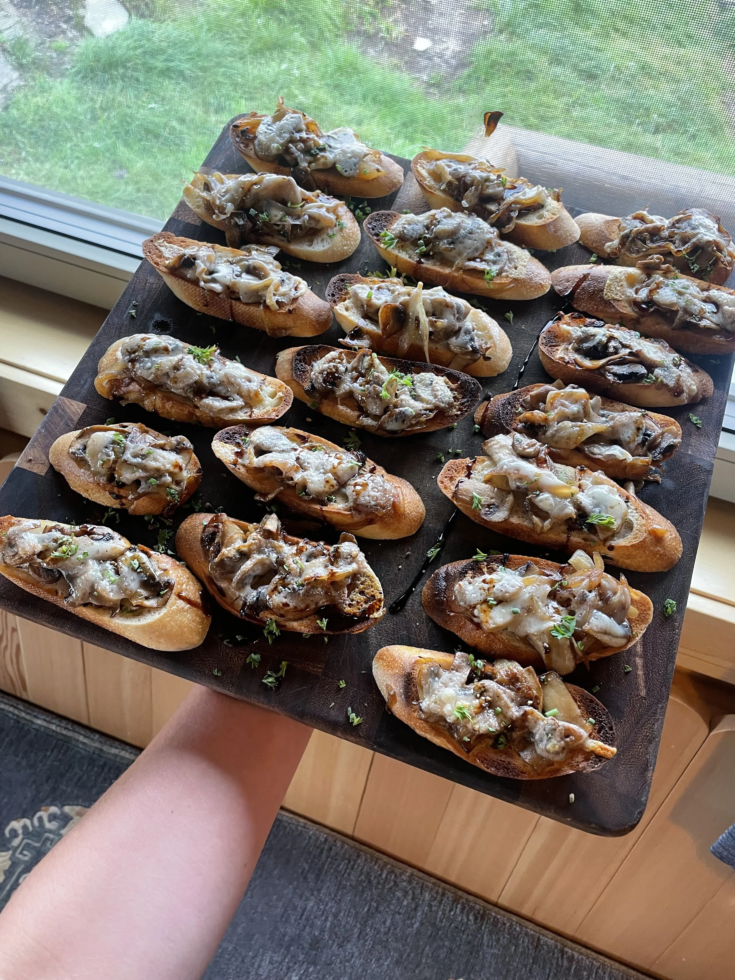 A hand holding a black tray with toasted bread slices topped with cooked mushrooms, melted cheese, and herbs, set near a window with a view of green grass outside prepared by a Revelstoke private chef.
