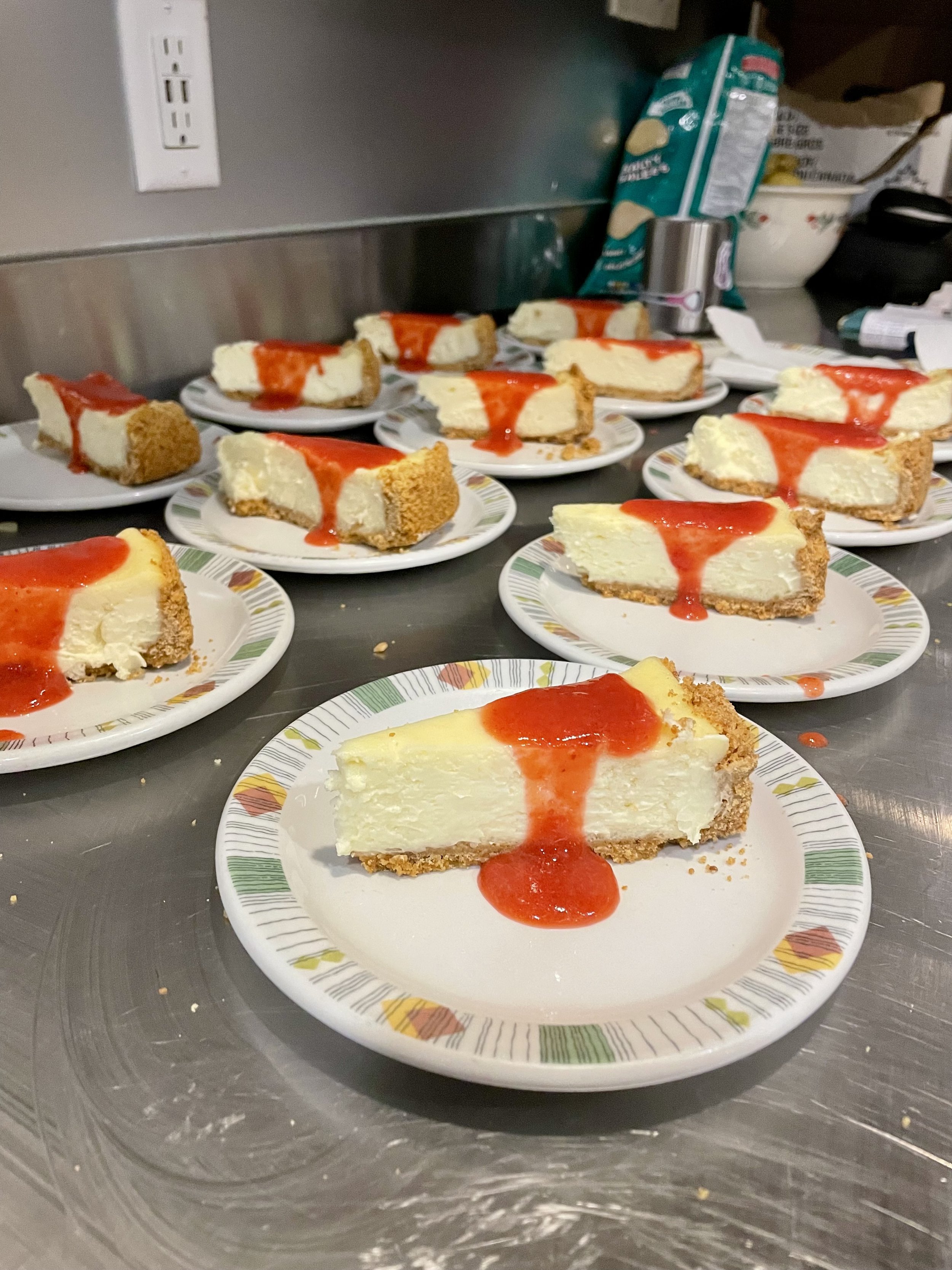 Slices of cheesecake with graham cracker crust and strawberry topping on paper plates, arranged on a stainless steel surface in a kitchen setting prepared by a Revelstoke private chef.
