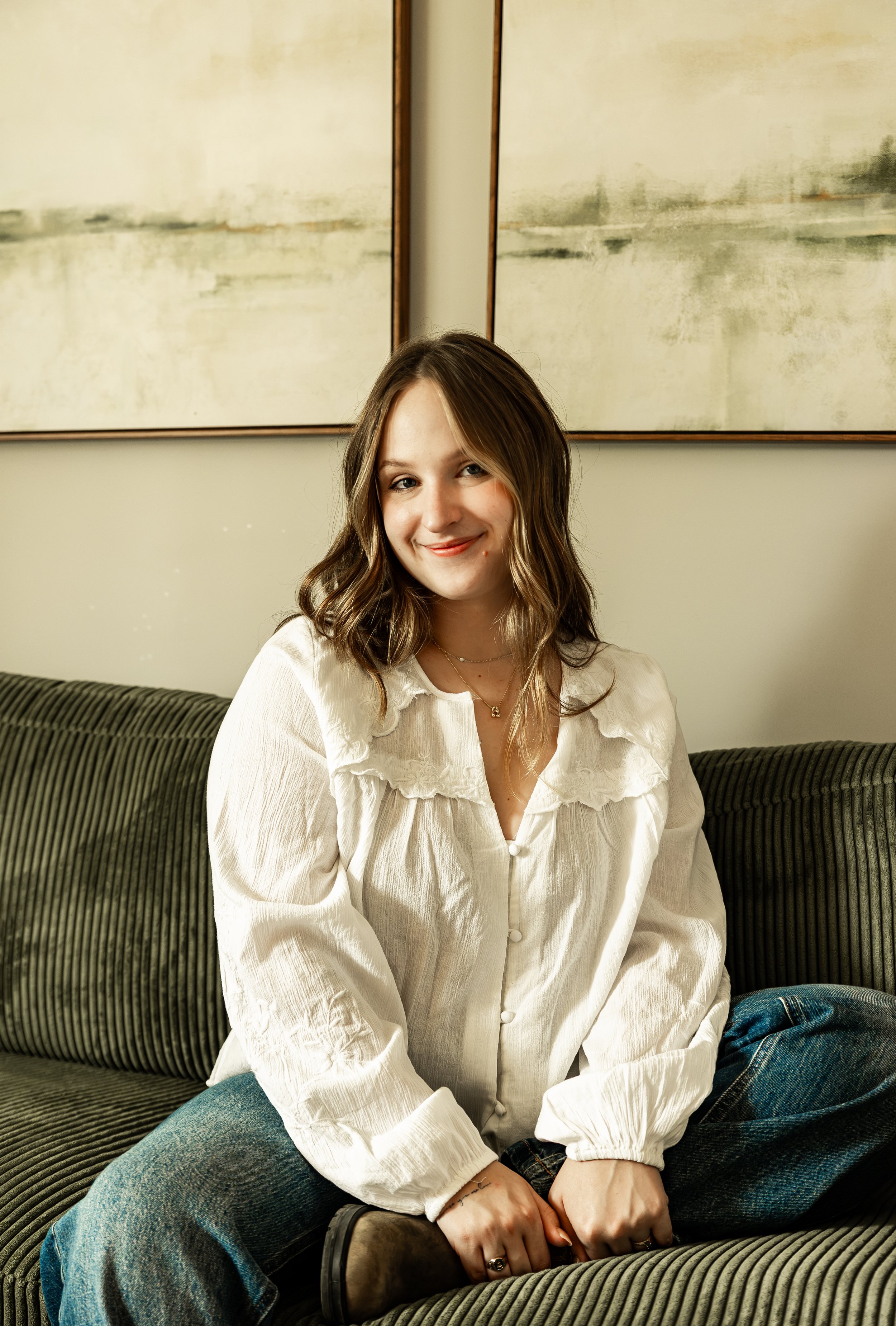 A young woman with wavy brown hair sitting on a green upholstered sofa in front of a beige wall with three abstract paintings. She is wearing a white blouse, blue jeans, and brown boots, and is smiling at the camera.