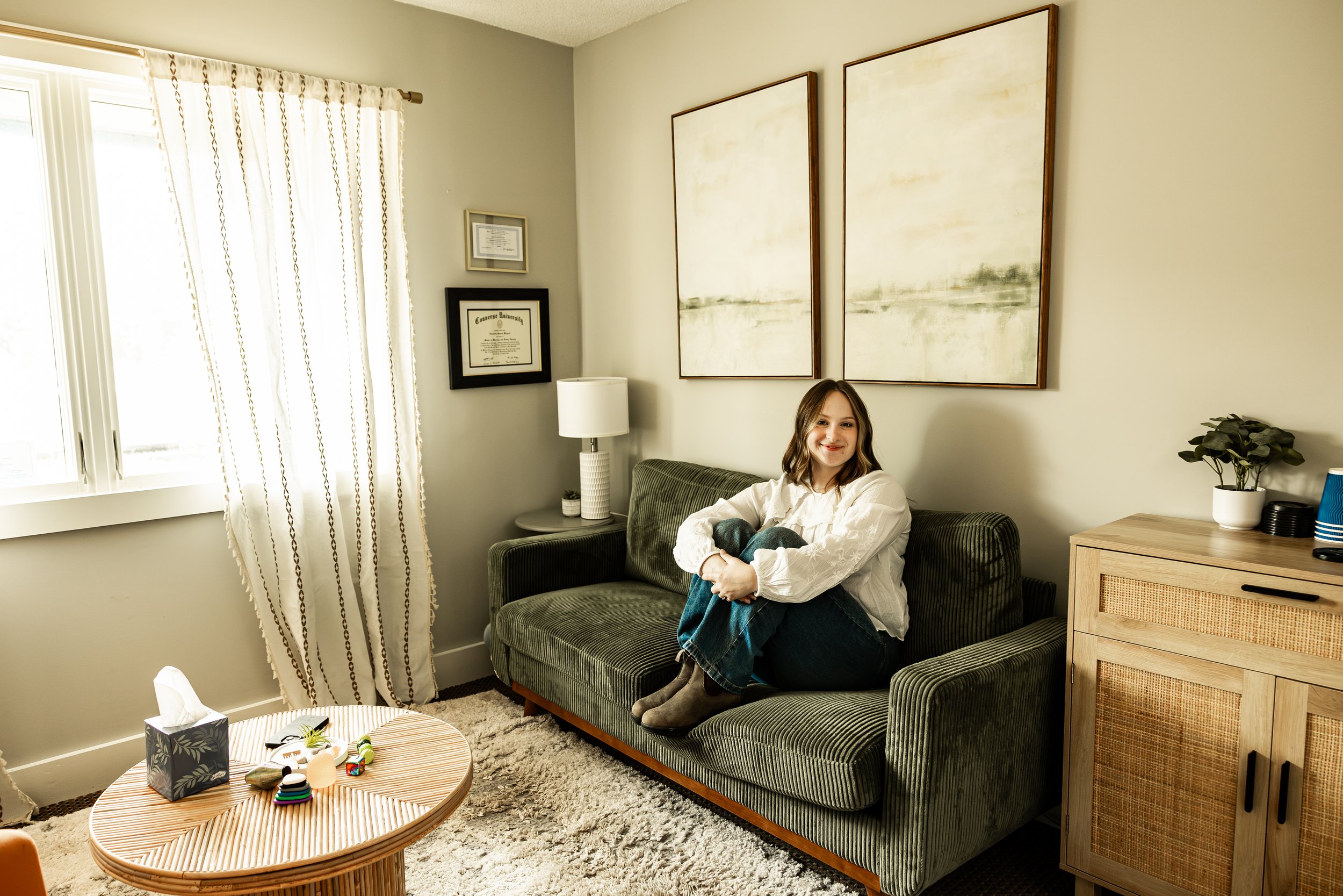 A woman sitting on a green sofa in a brightly lit living room. The room has beige walls, two large abstract paintings, a window with white curtains, a wooden side table with a white lamp, another framed certificate, a small potted plant, and a wooden cabinet. A round coffee table with assorted items is in the foreground, and a plush gray rug covers the floor.