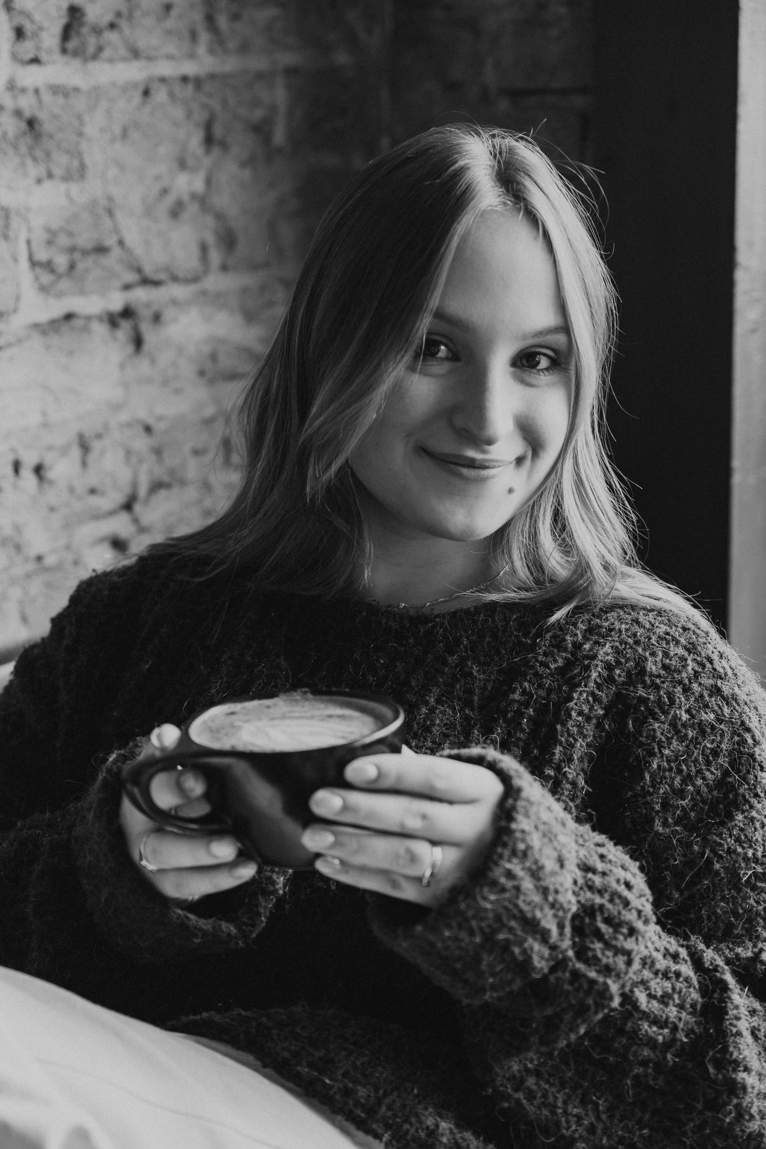 A young woman with long hair smiling and holding a mug, sitting against a brick wall.