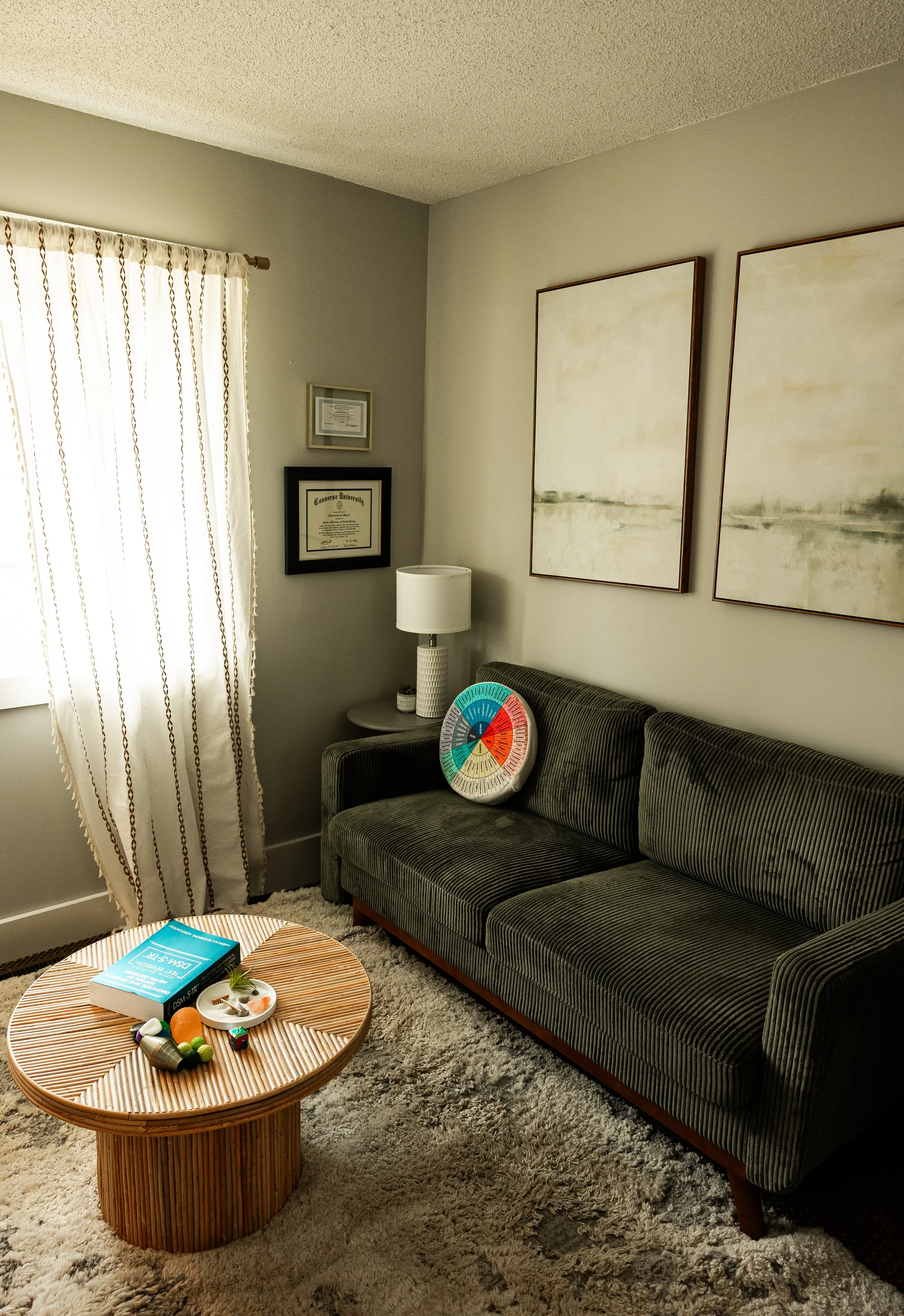 Living room with a striped green couch, a round wooden coffee table, a beige shag rug, wall art, a white lamp, and a window with white curtains.