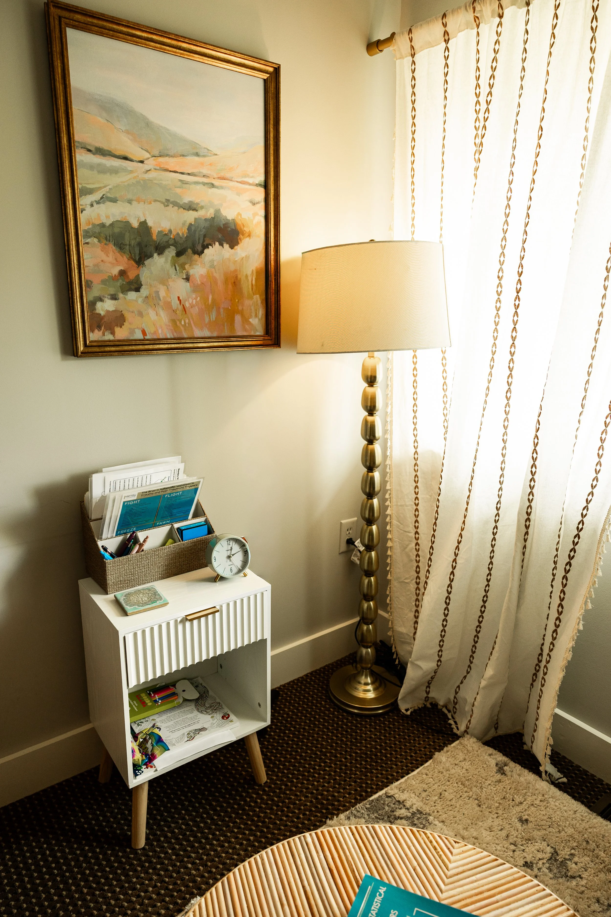 A cozy corner of a room featuring a framed landscape painting, a tall beige and gold floor lamp, a white miniature bookshelf with various items including a clock, a coloring book, and some art supplies, and a window with cream-colored curtains with a