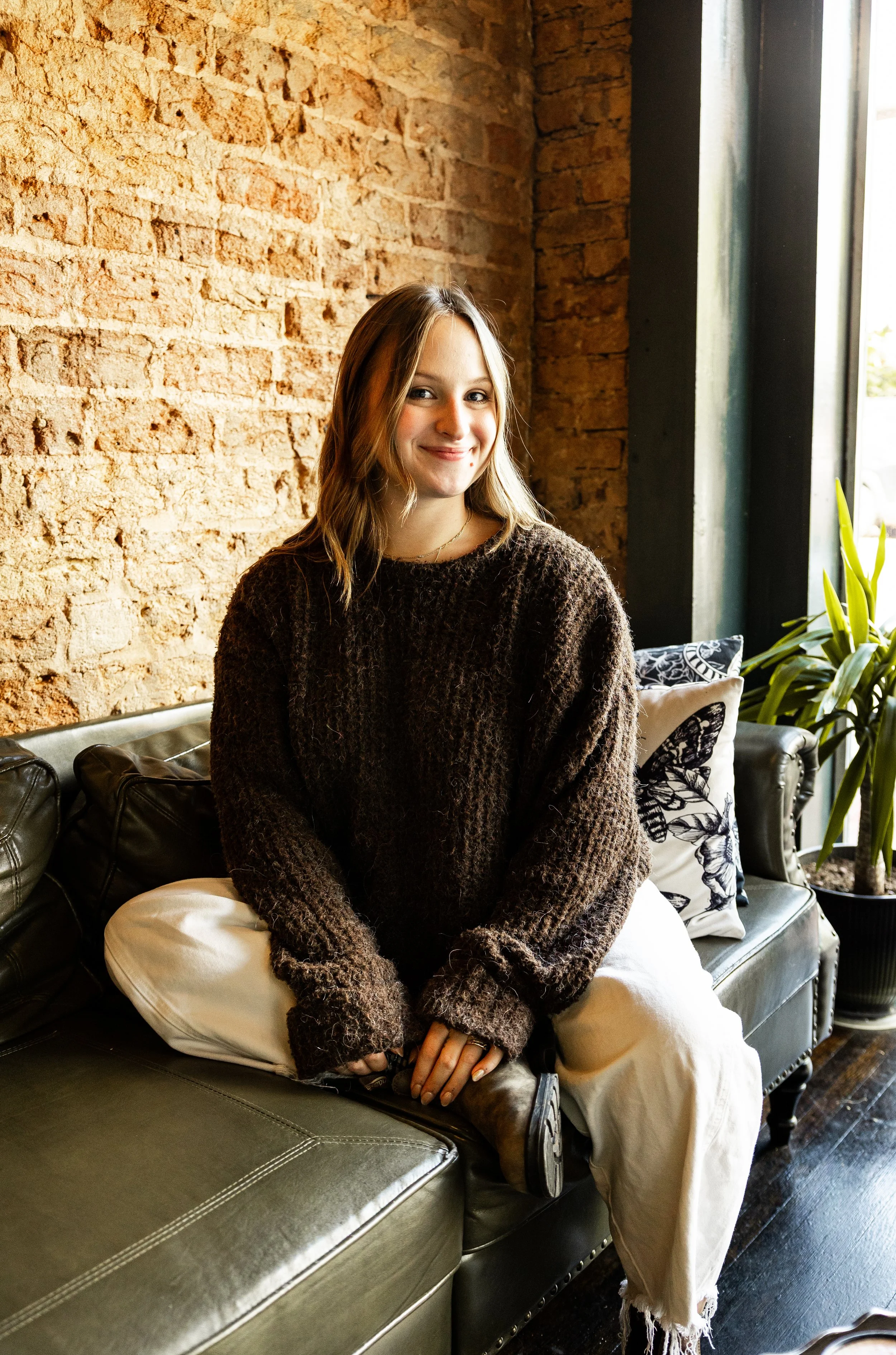 A young woman with long hair, wearing a brown fuzzy sweater and white pants, sitting on a dark leather couch with a decorative pillow, in a room with a brick wall and large window with sunlight.