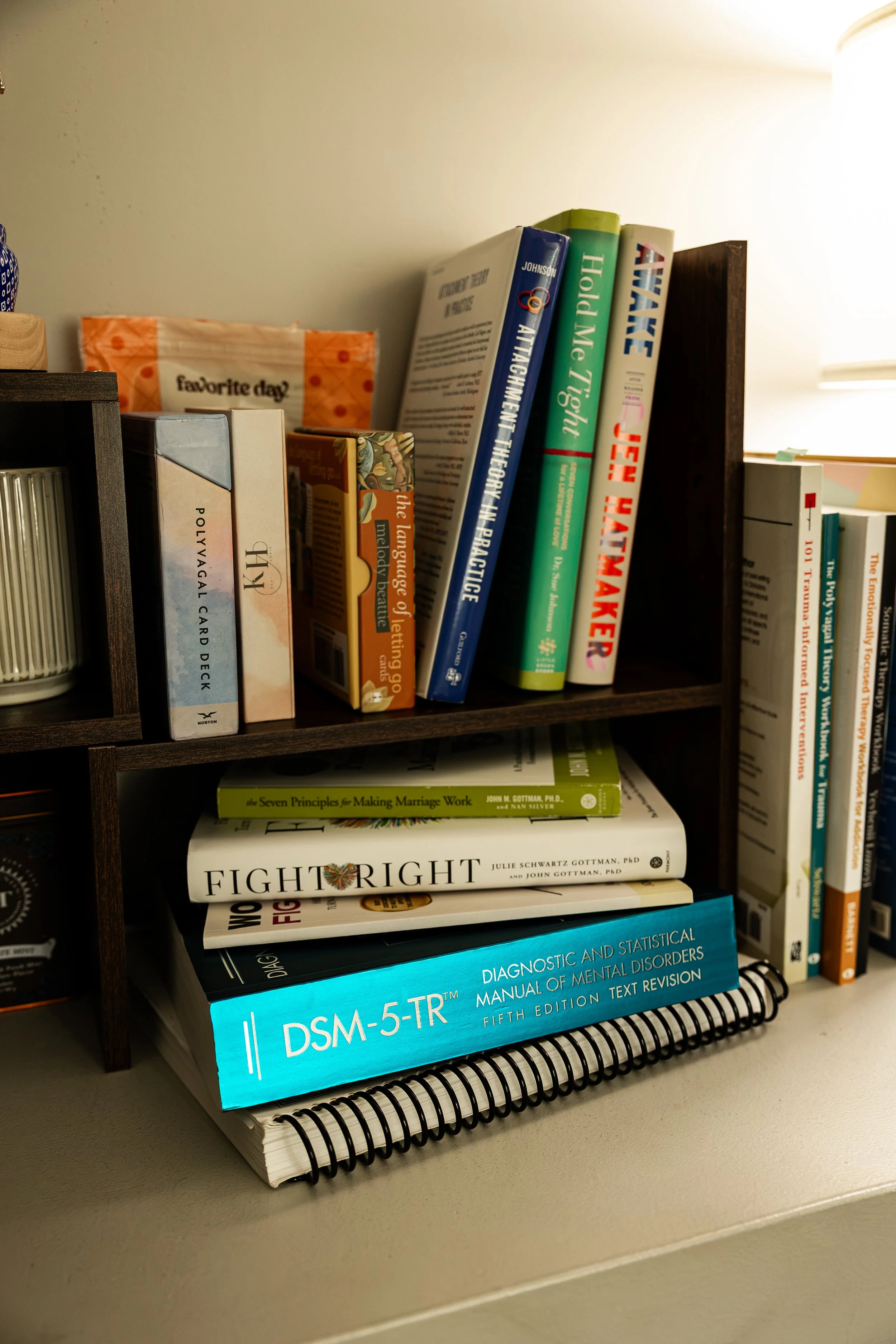 Bookshelf with various books on psychology, mental health, and self-help, including titles like 'The Language of Feeling,' 'Fight or Flight,' and 'DSM-5-TR,' with a spiral notebook underneath.