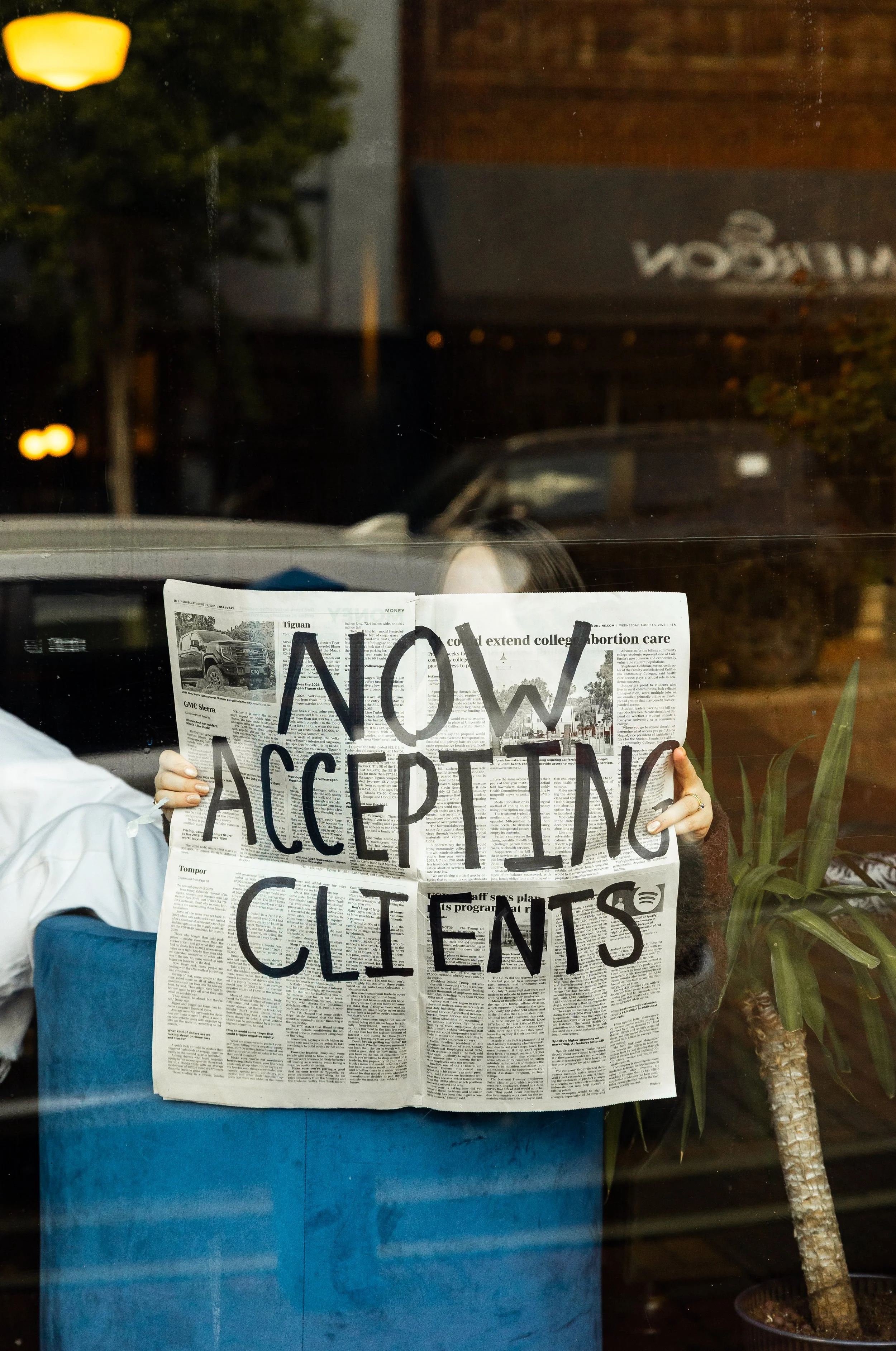Person holding a newspaper with the words 'NOW ACCEPTING CLIENTS' written in large black letters, with a reflection of a truck and some trees outside a window.