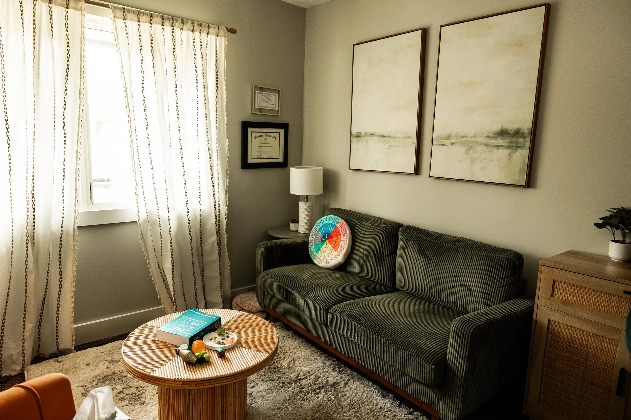 A cozy living room with a window and cream-colored curtains on the left, and artwork hanging on the wall on the right. There is a gray striped sofa with a white pillow, next to a wooden side table. A white table lamp is on the side table, and a small