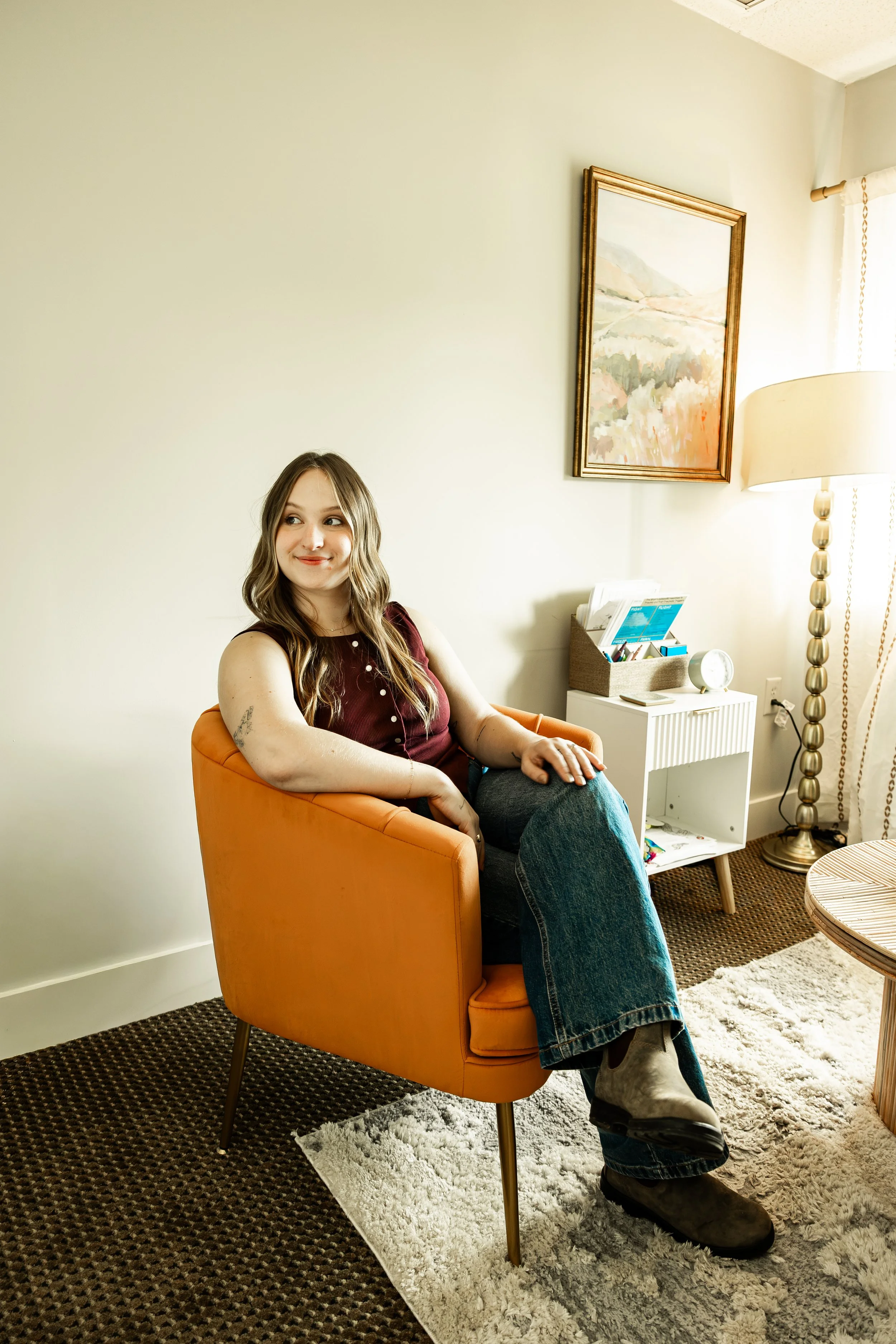 A woman with long wavy hair sitting in an orange armchair in a living room with a cream-colored wall, a framed painting, a white side table with magazines and a clock, a tall decorative floor lamp, and a window with curtains. She's smiling and looking to her left.