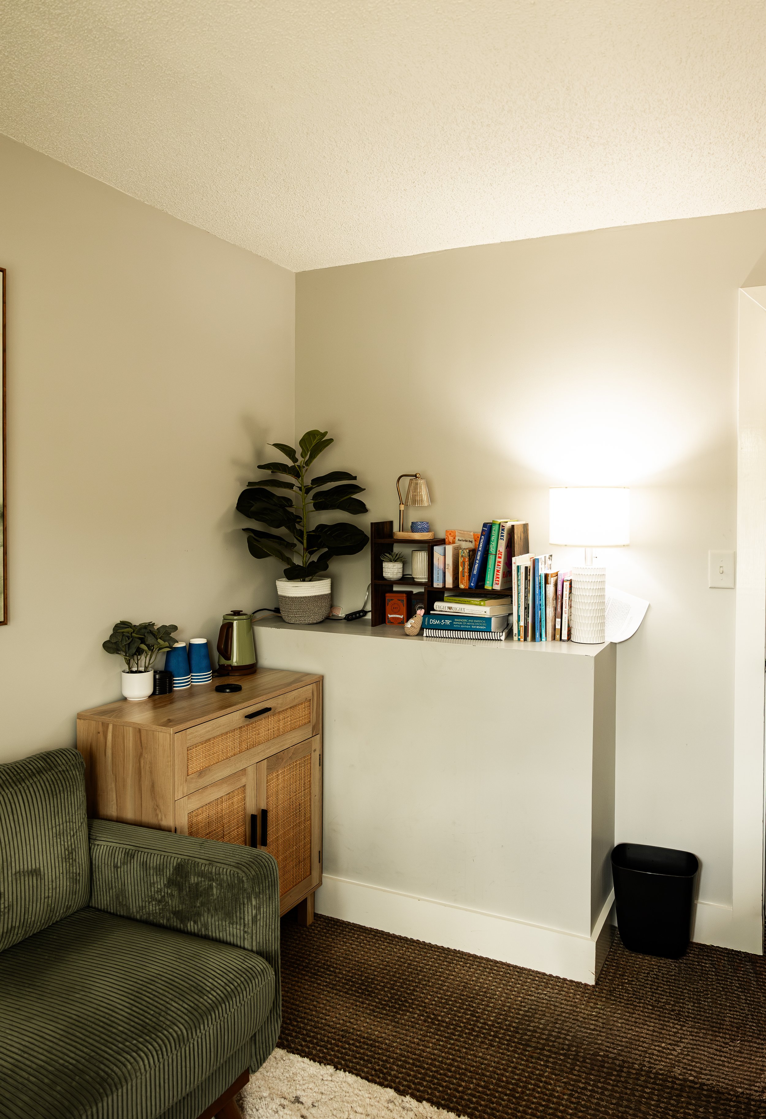 Living room corner with a green armchair, a wooden cabinet, and a wall with a bookshelf, plants, and a lamp