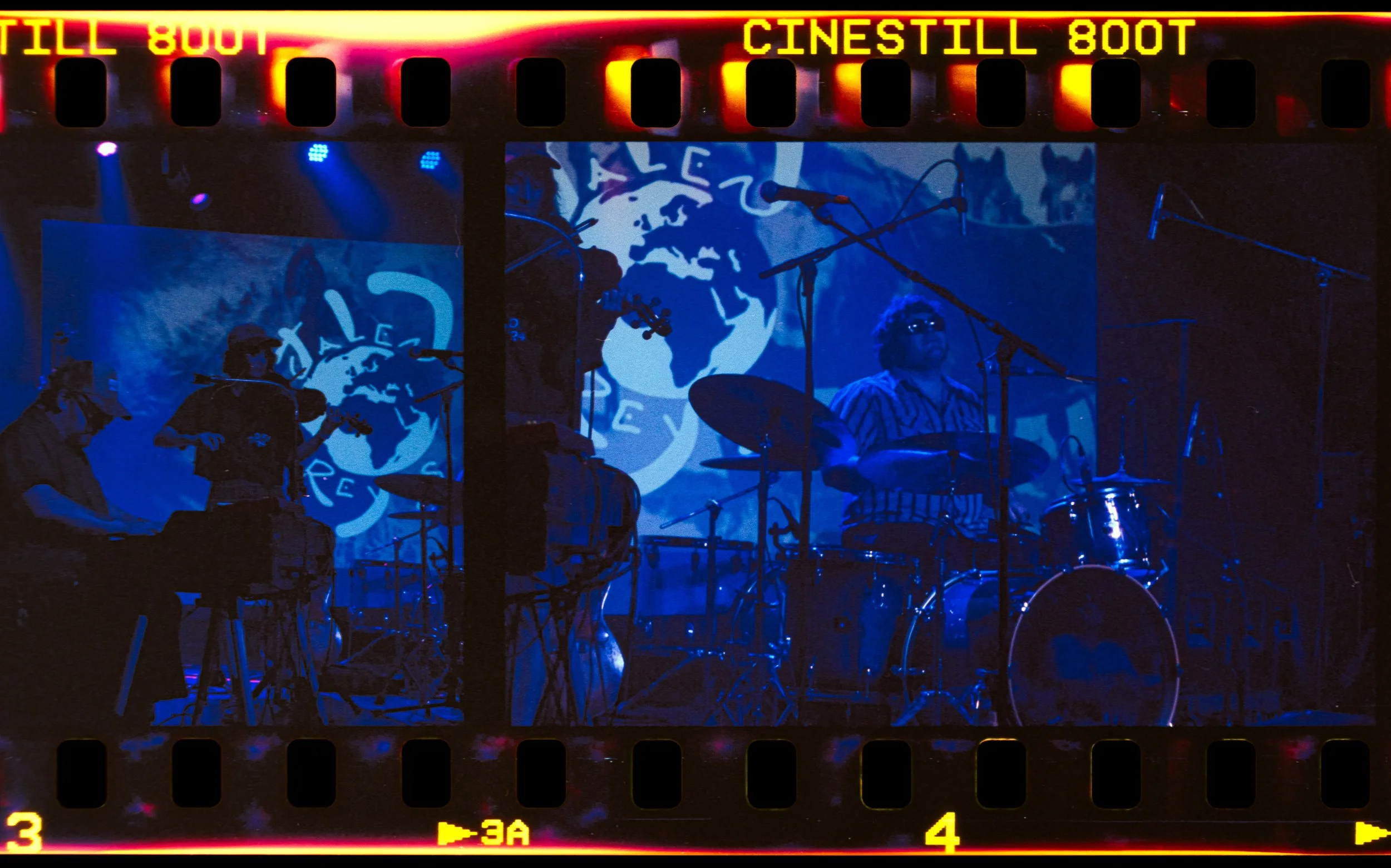 Musicians performing on stage with blue lighting and a backdrop displaying a logo featuring a globe and the word "AL". A drummer is visible in the foreground, and other band members are playing instruments.