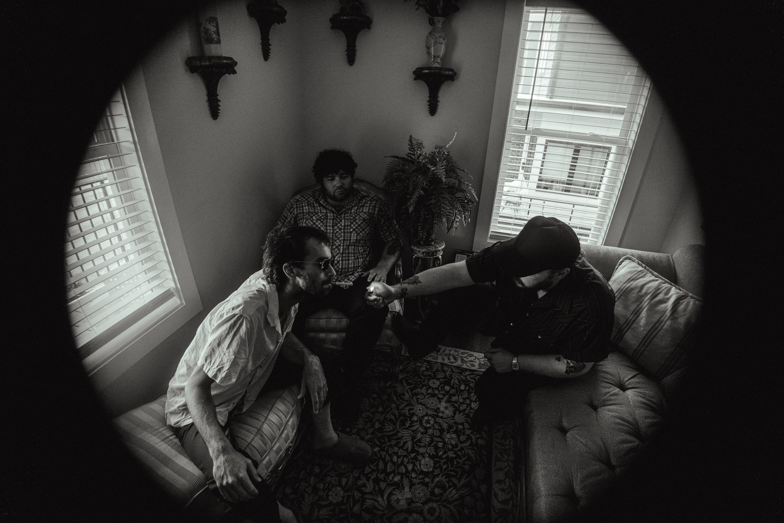 A black and white photo of a living room scene viewed through a circular peephole. Four people are sitting and lying on a striped sofa and a floral-patterned armchair. One person is smoking a cigarette, another is leaning against a large potted fern plant, and a third is reaching out to the person smoking. The room has two windows with blinds, three decorative wall sconces, and ornate vases on the wall.