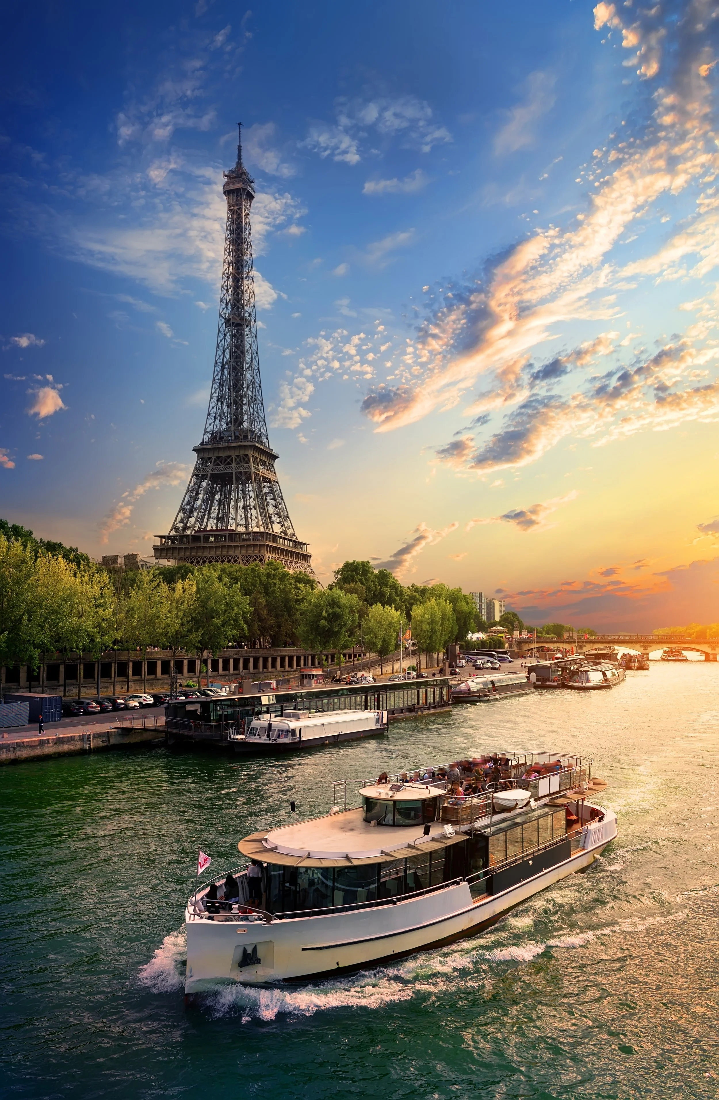 The Eiffel Tower along the Seine River in Paris, France, with a luxury river cruise boat on the water and a sunset sky with scattered clouds.
