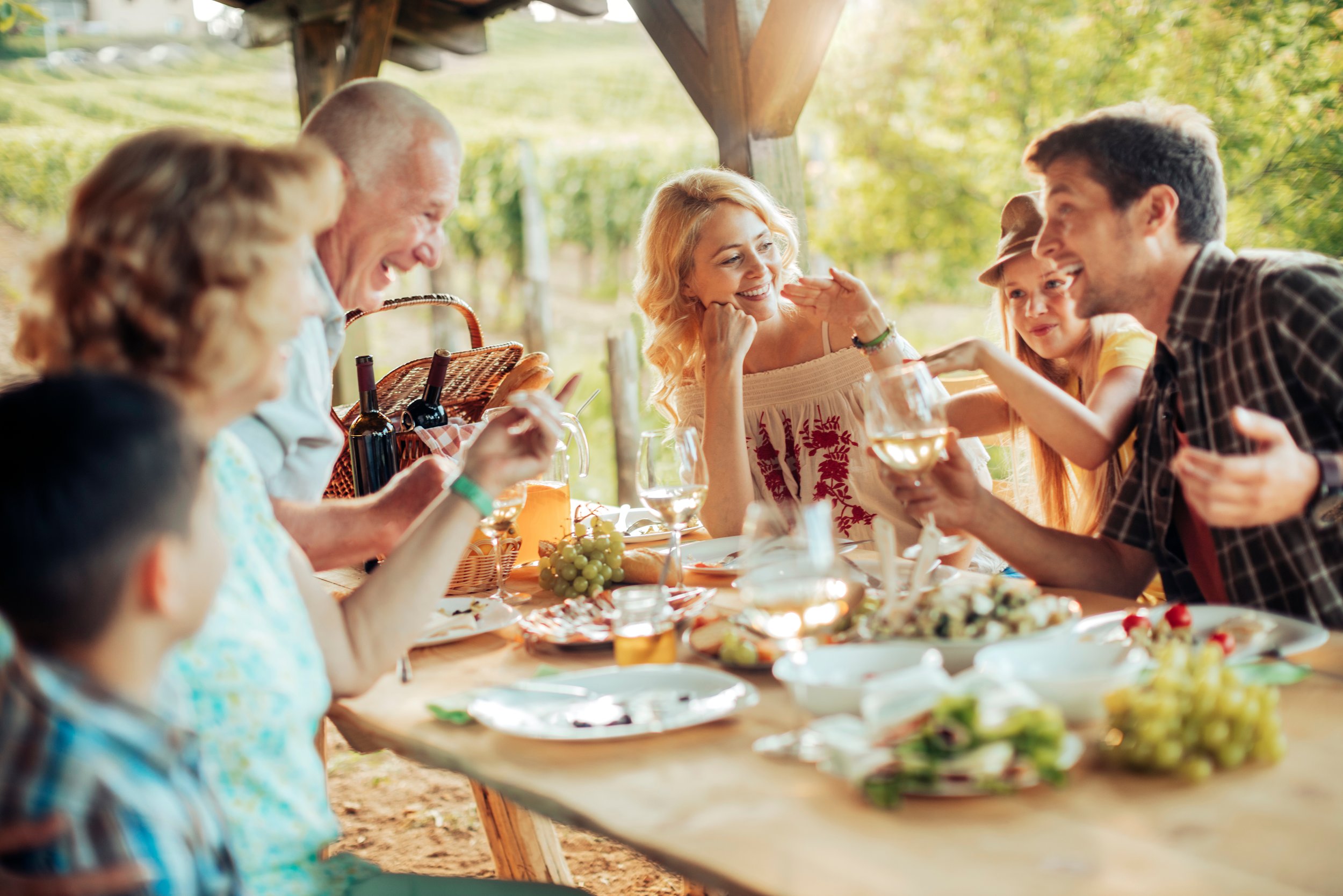 A group of people sitting at a table outdoors, enjoying a meal and drinks, with trees and sunlight in the background, smiling and engaging in conversation.