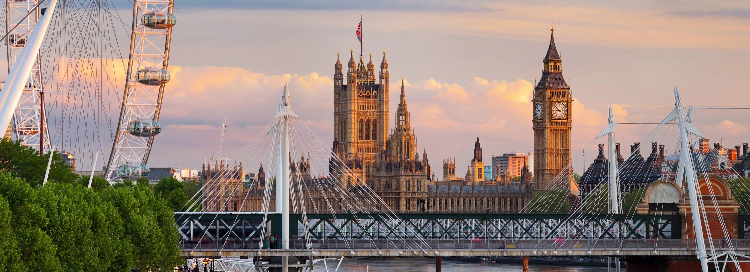 View of the London skyline at sunset, featuring the London Eye, the Houses of Parliament, and Big Ben, with a partly cloudy sky in the background.