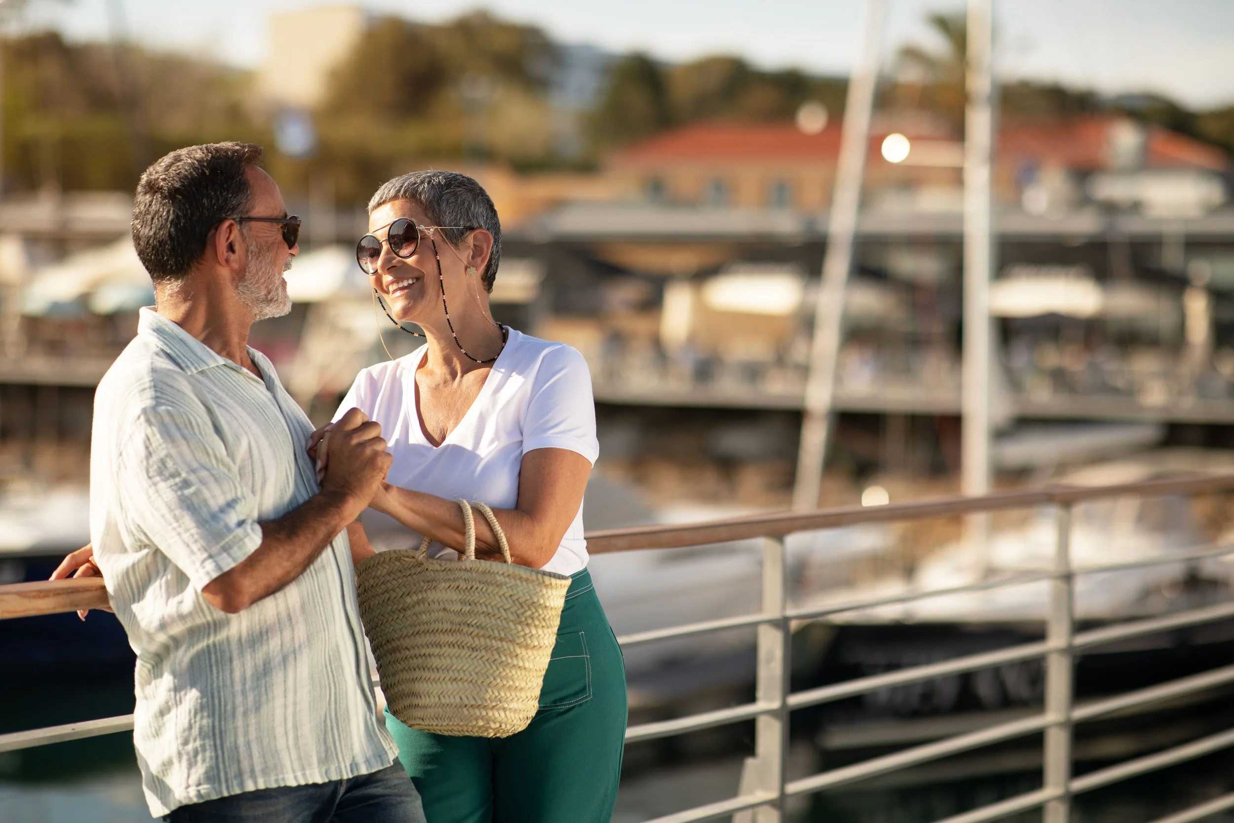 A happy older couple with short gray hair and sunglasses, smiling and holding hands on a waterfront dock with boats and buildings in the background.