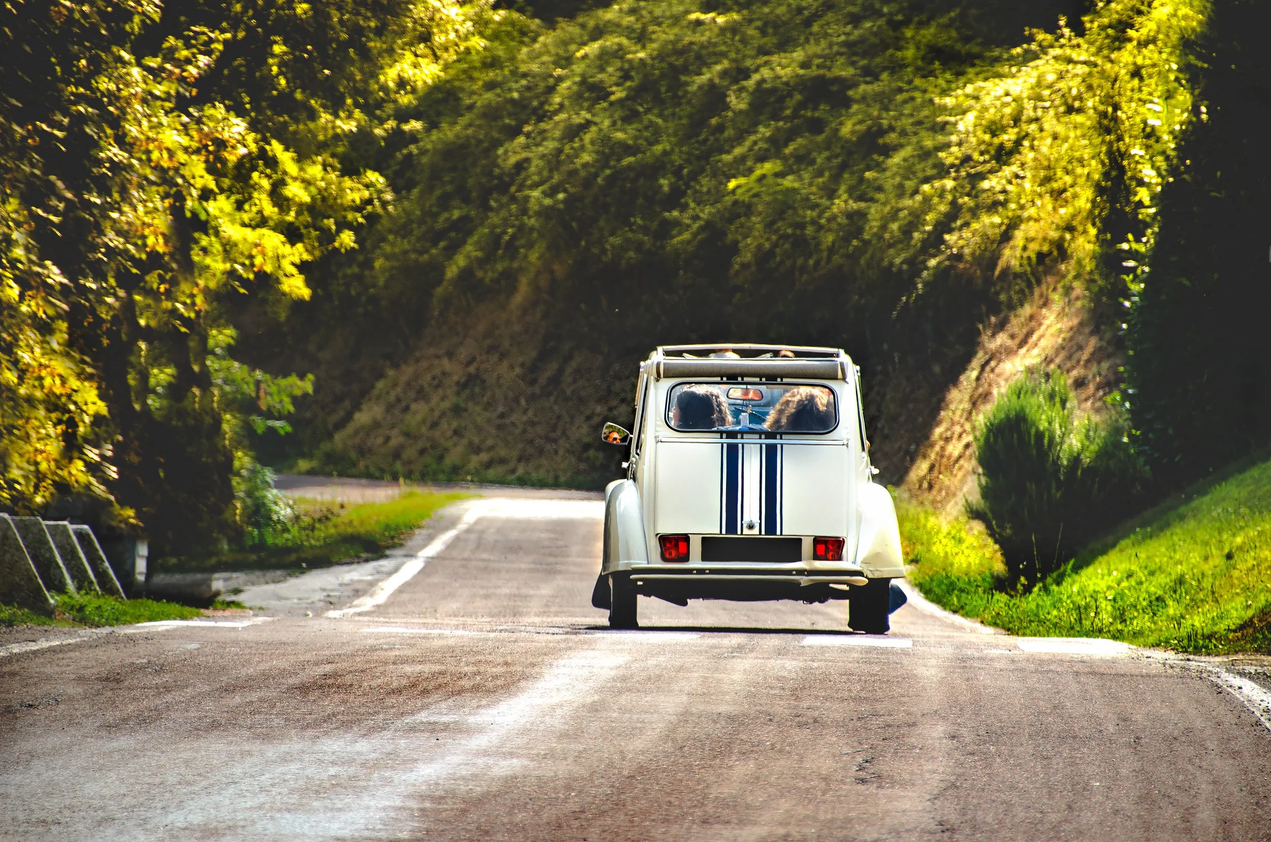 A white vintage French Citroen car with black racing stripes driving on a road in the French countryside surrounded by lush green trees.