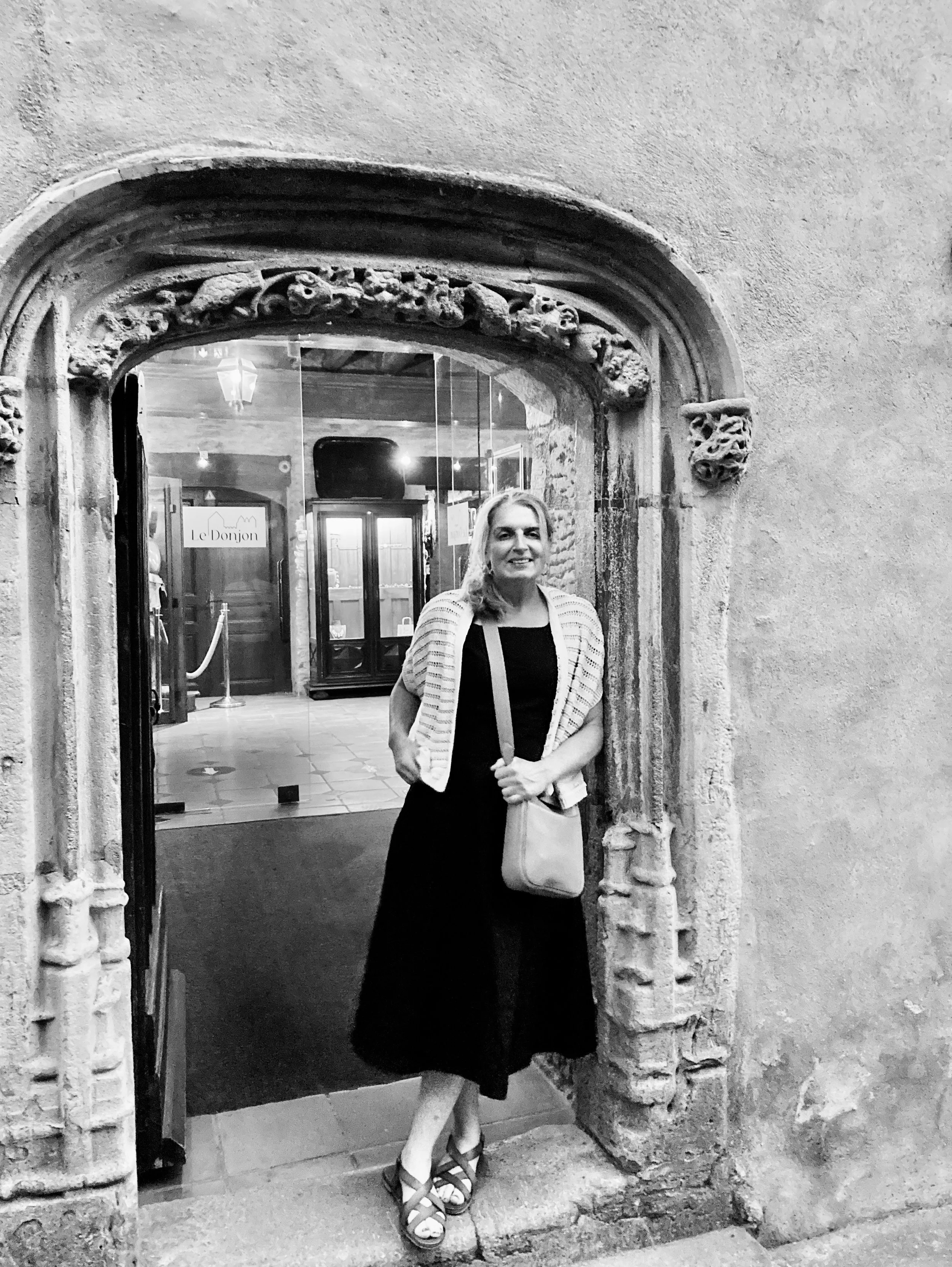 A woman standing in front of an ornate stone doorway in Carcassonne, France with a glass door behind her. She is smiling, wearing a black dress, a light-colored checkered shawl, and sandals, holding a beige purse and a tote bag.