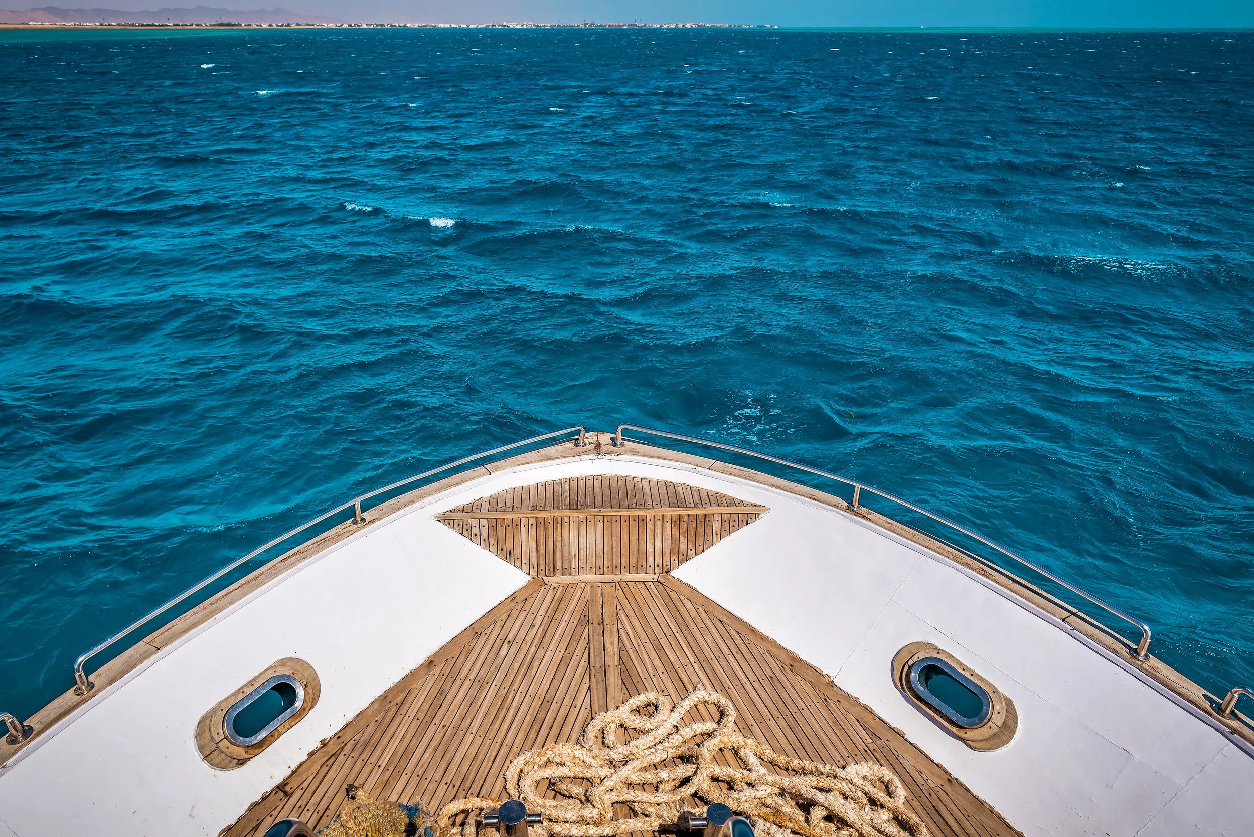 View from the front of a boat with a wooden deck, looking out onto blue ocean water and a distant shoreline with mountains.
