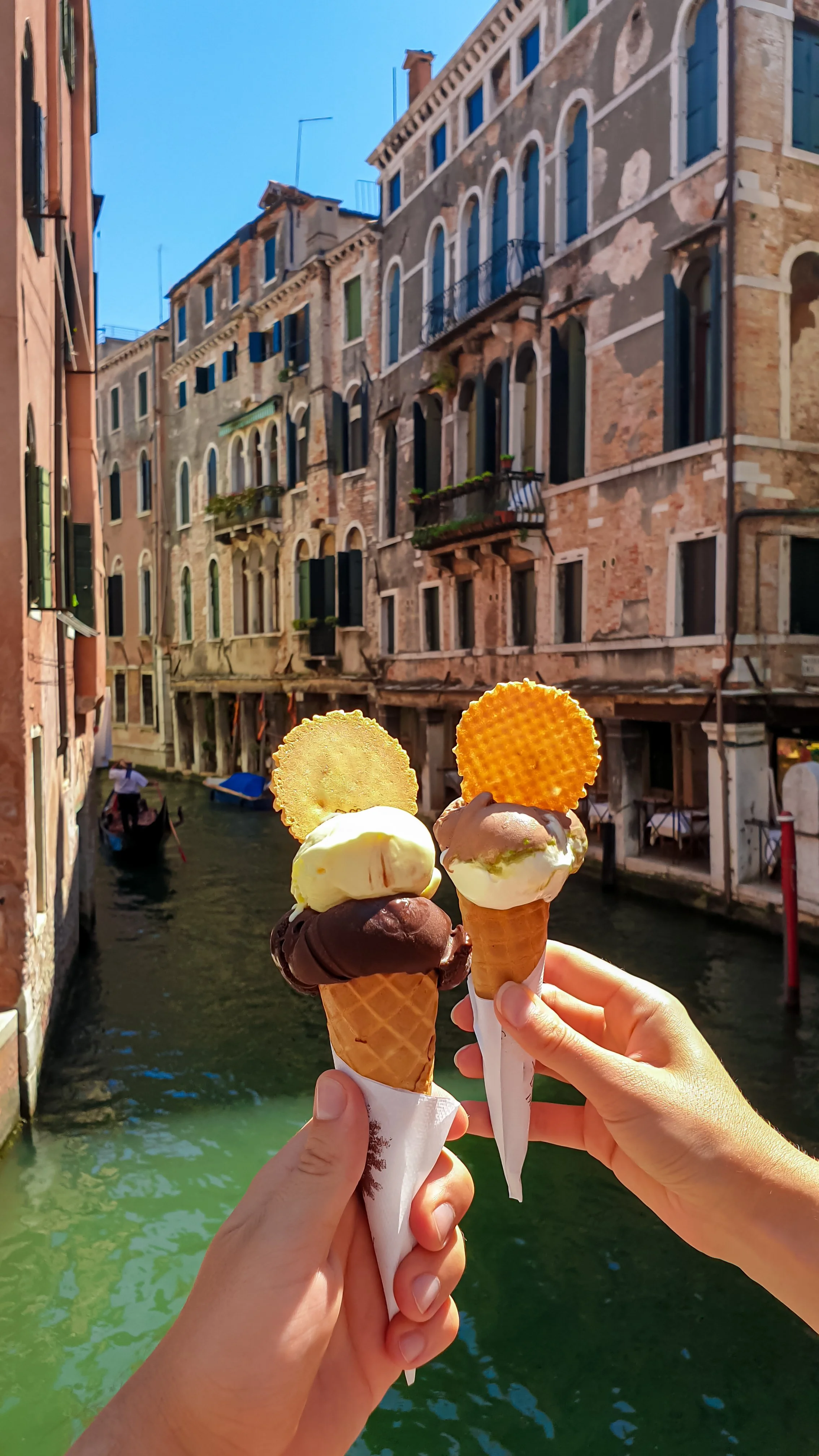 Two hands holding three ice cream cones with various flavors in front of a canal with old brick buildings and a boat in Venice, Italy.