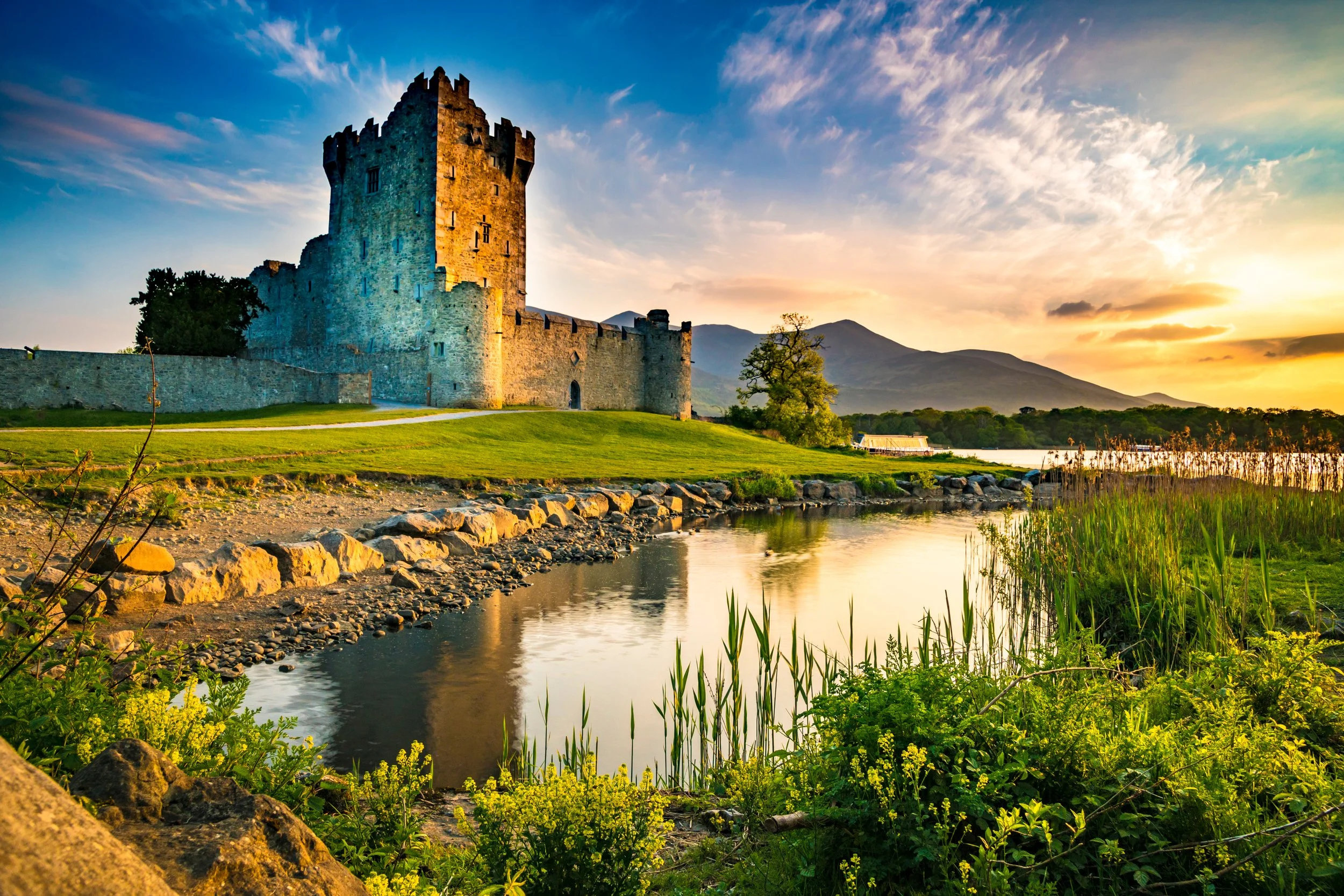 Ross castle in Kilarney, Ireland on a grassy hill next to a river, with mountains and a colorful sunset sky in the background.