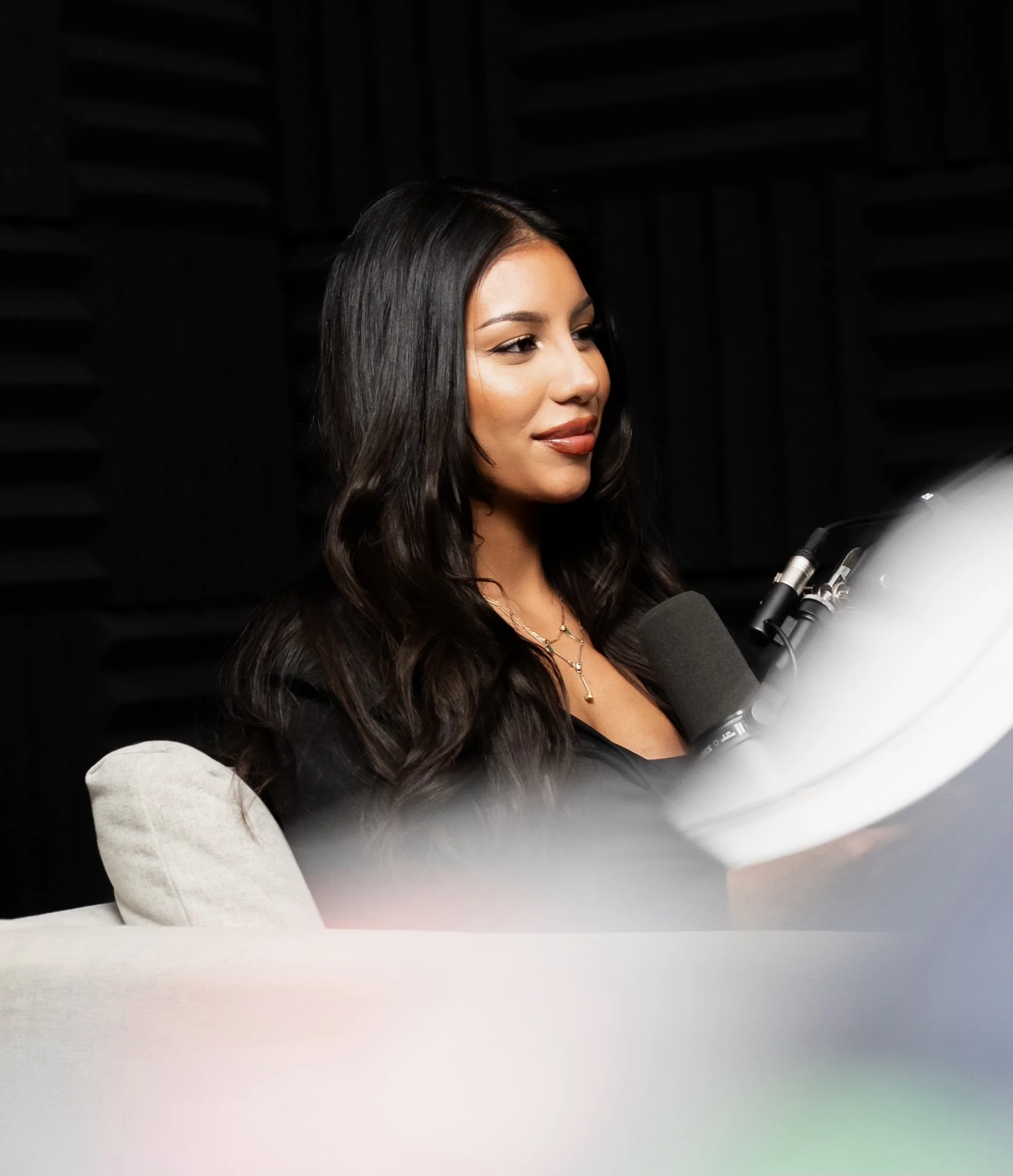 A woman with long dark hair sitting at a white desk, looking at a computer screen, with a microphone in front of her, in a recording studio.