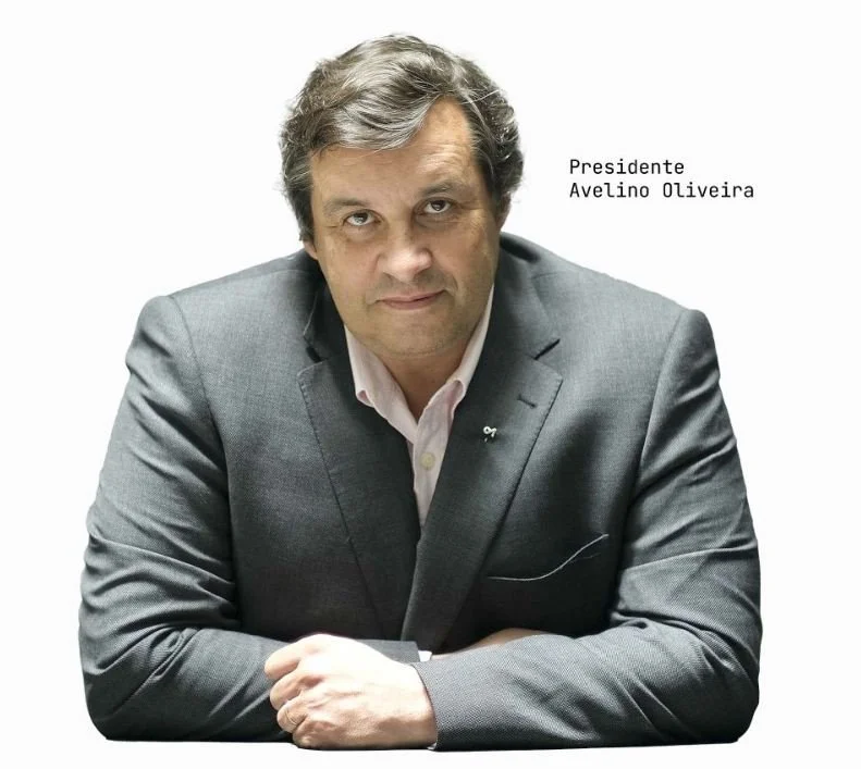 Homem de terno cinza, com cabelo castanho e grisalho, sentado com braço cruzado, texto ao lado indicando que é o presidente Avelino Oliveira.