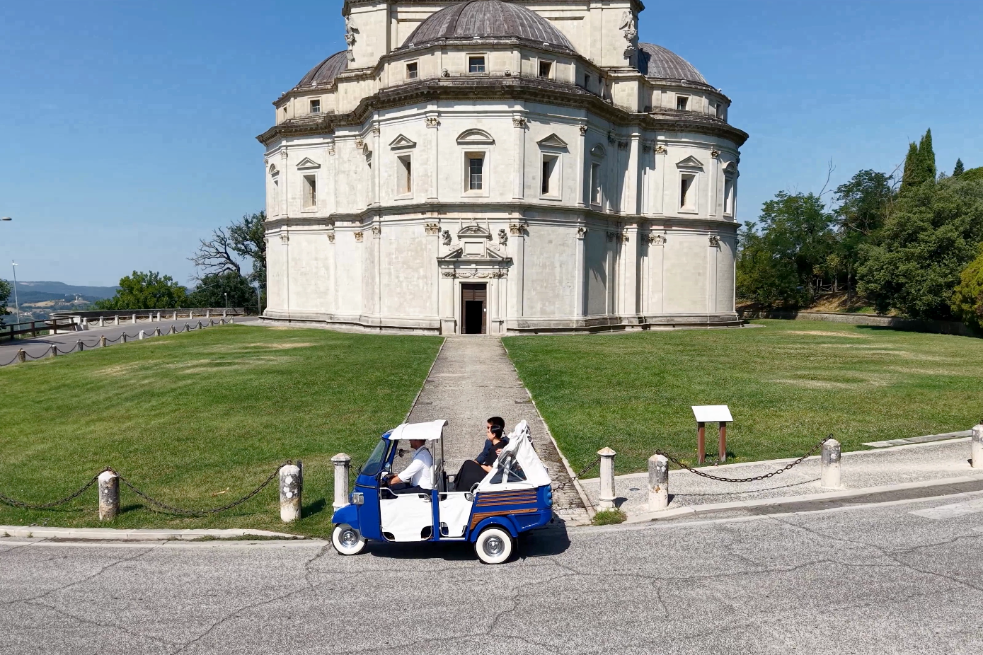 A white and blue electric cart with two men riding past a historic, circular stone building with domed roofs, set on a manicured lawn on a sunny day.