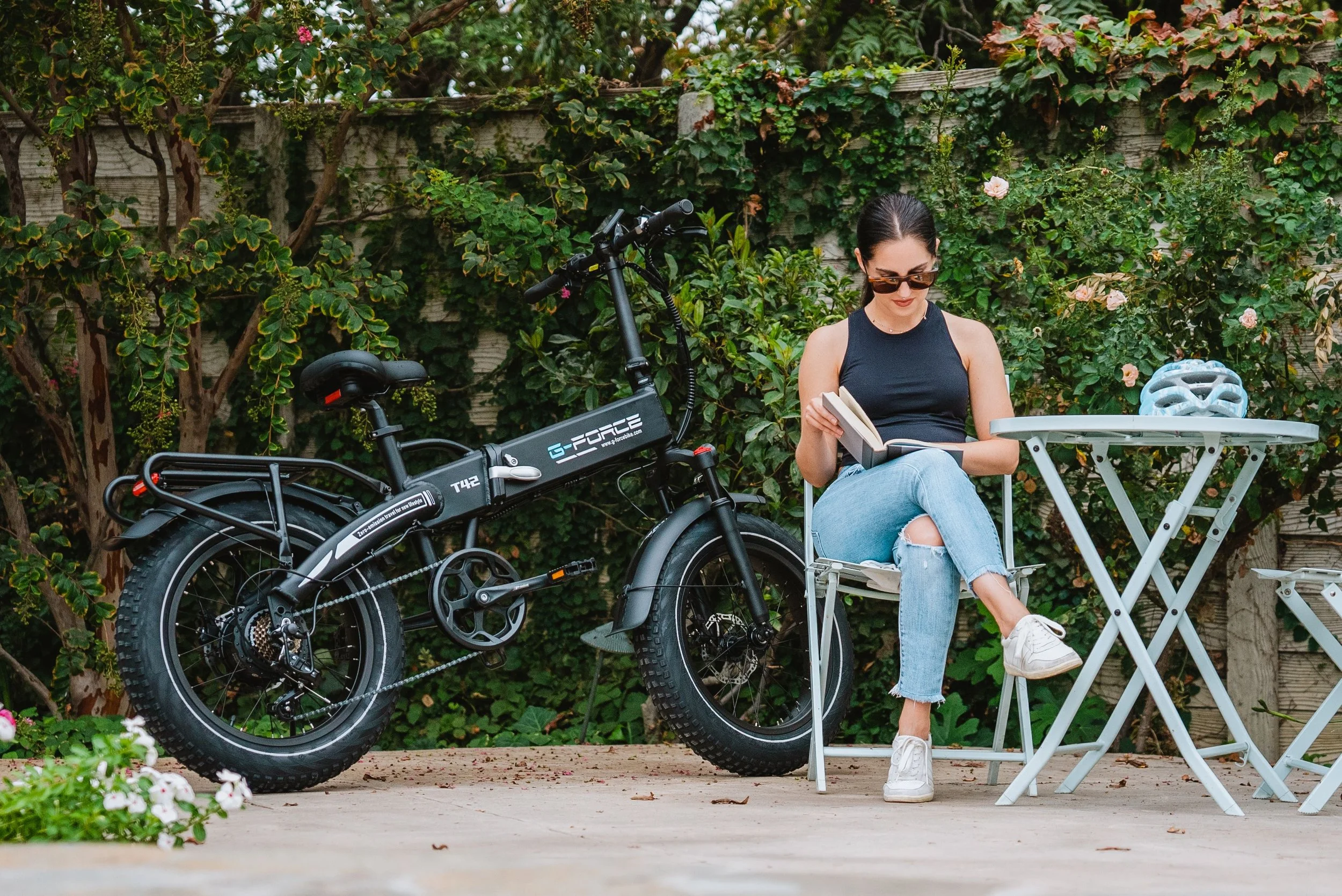 A woman sitting outdoors at a small table with a bicycle parked beside her, reading a book, with a bicycle helmet on the table and a lush green hedge in the background.