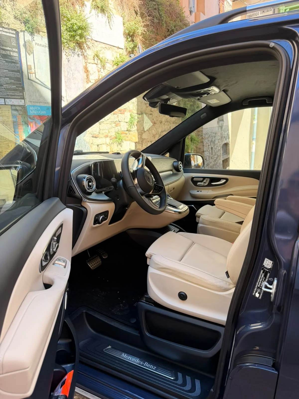 Interior view of a luxury Mercedes-Benz vehicle with beige seats, black dashboard, and steering wheel, parked beside a stone wall and greenery visible through the window.