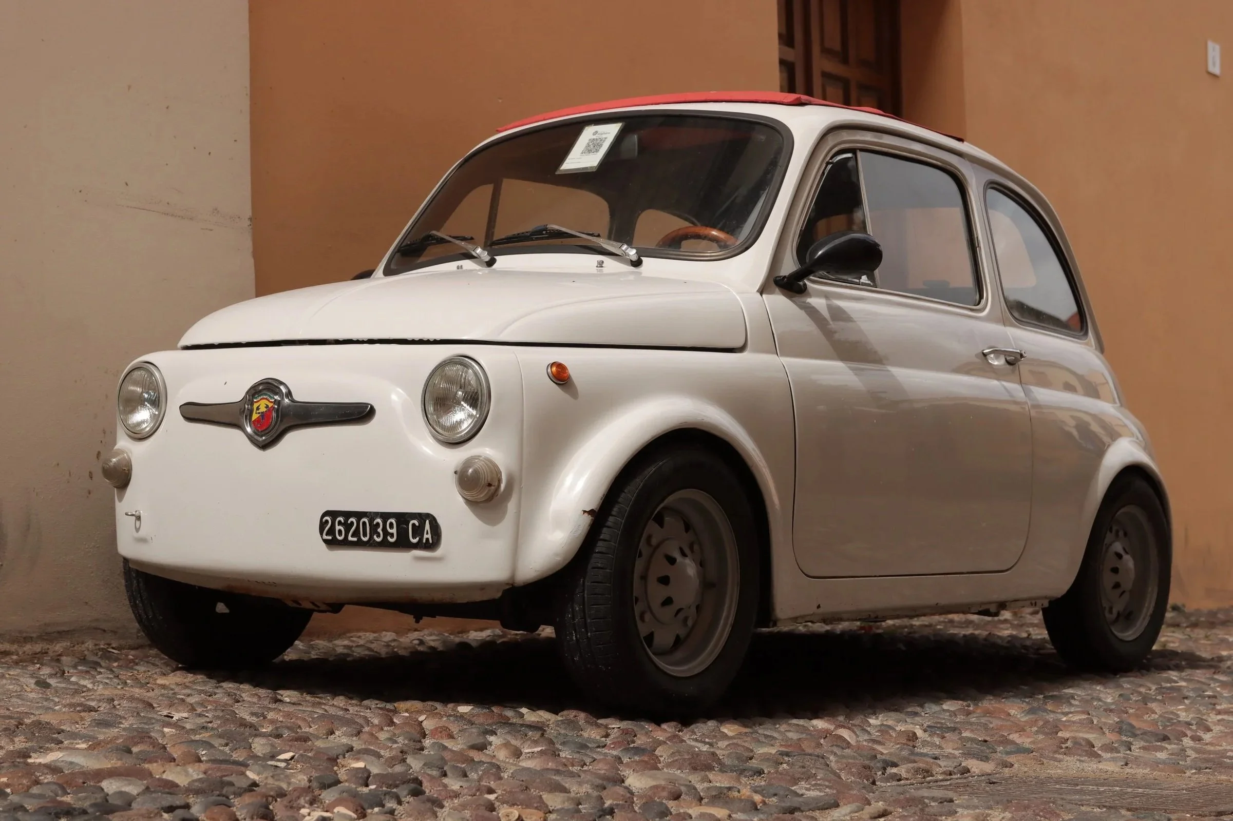 A vintage white microcar with a red roof, parked on a cobblestone street against a beige wall. The front features round headlights, a black emblem with a yellow and red design, and a black license plate that reads 262039 CA. The car has black side mirrors and a QR code on the windshield.