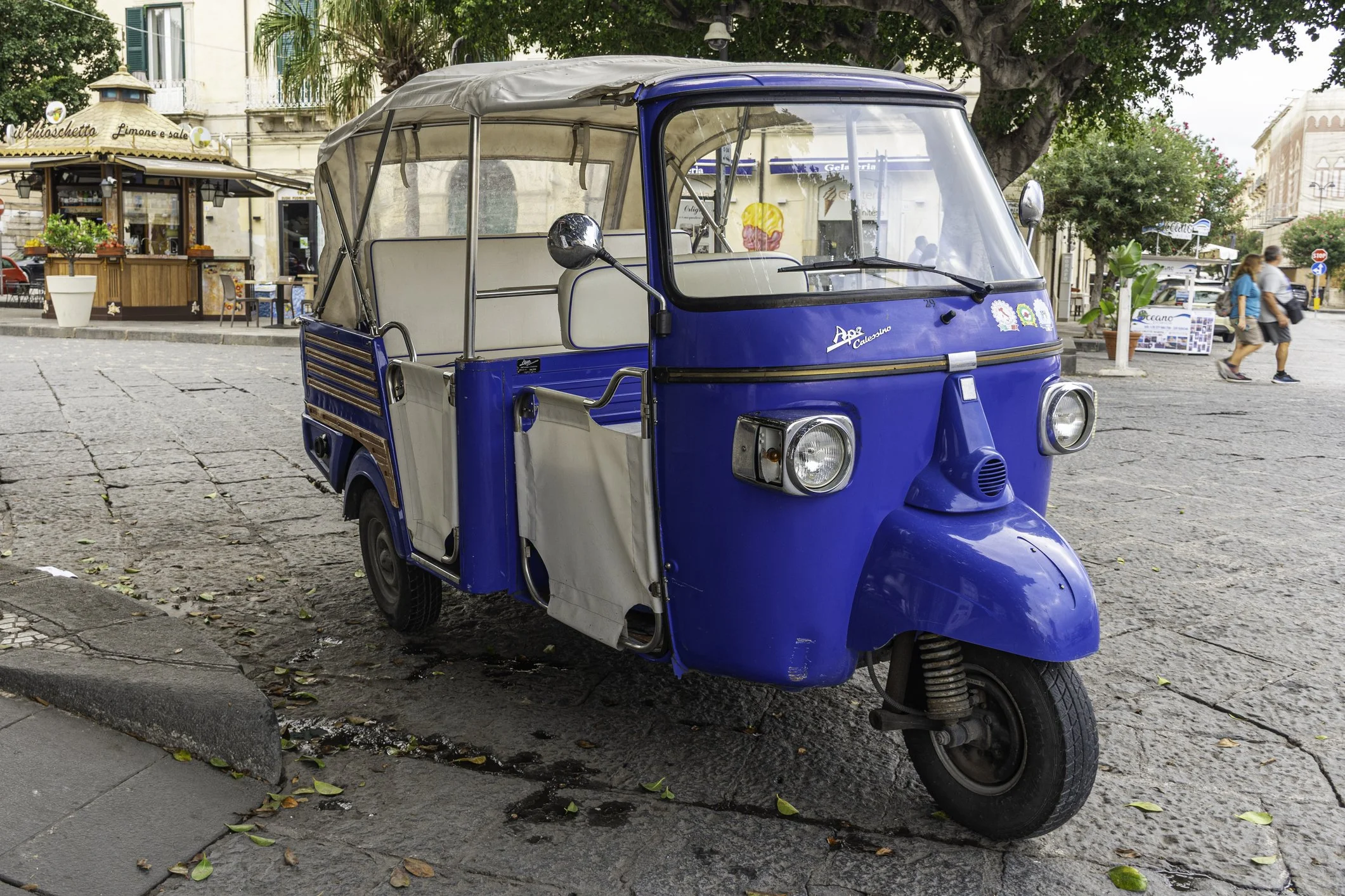 A blue electric tuk-tuk parked on a cobblestone street in a European city. In the background, there are trees, a cafe, and pedestrians.