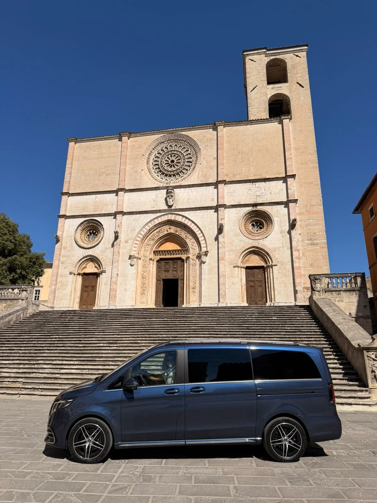 Facade of a historic church with stone stairs leading up to three ornate wooden doors, topped with a circular rose window, and a bell tower on the right side, under a clear blue sky. A gray minivan is parked in front of the stairs.