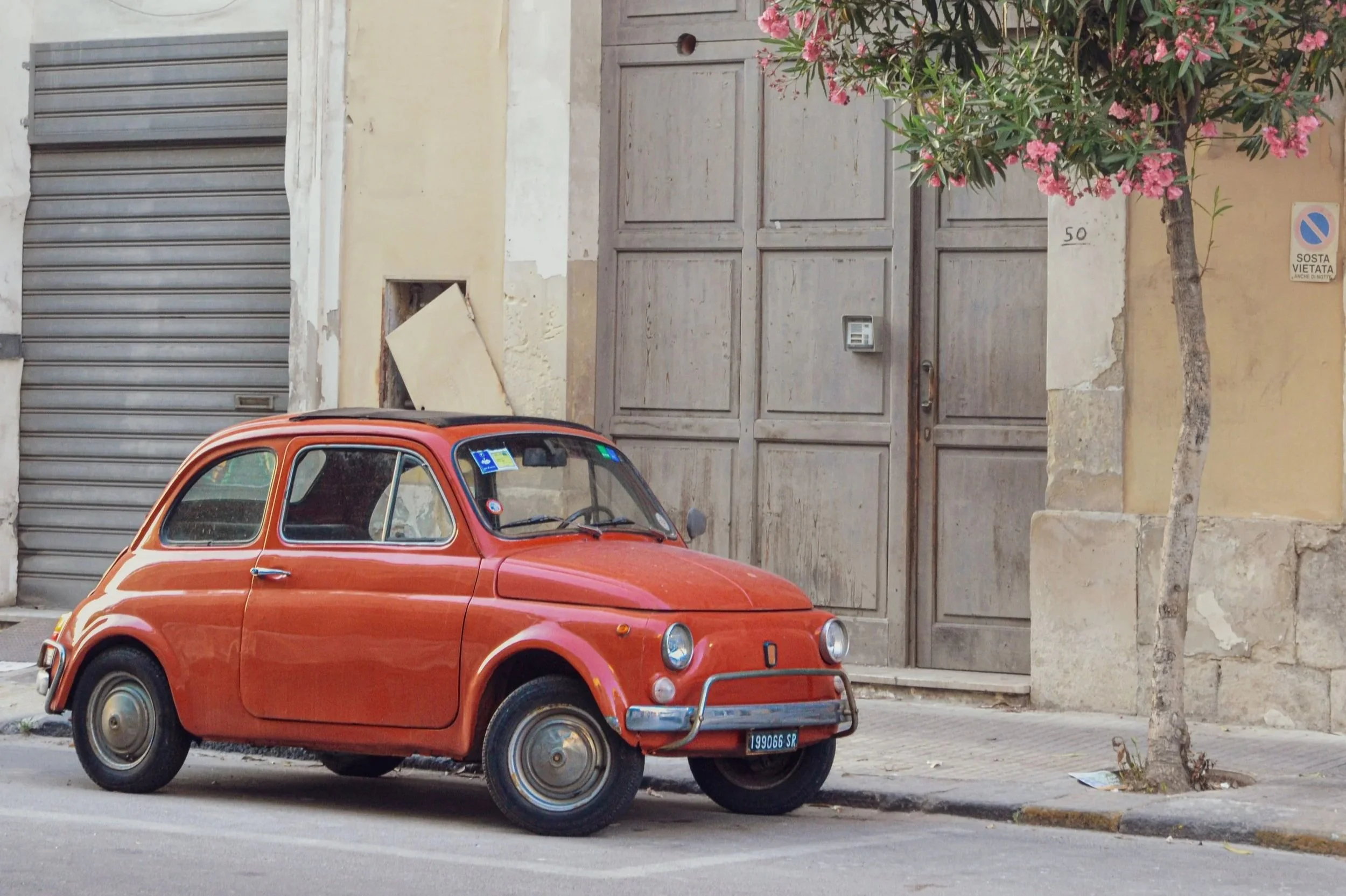 A vintage orange car parked on a paved street in front of a beige building with a closed garage door and a wooden gate. There is a pink flowering tree and a 'No Parking' sign on the wall.
