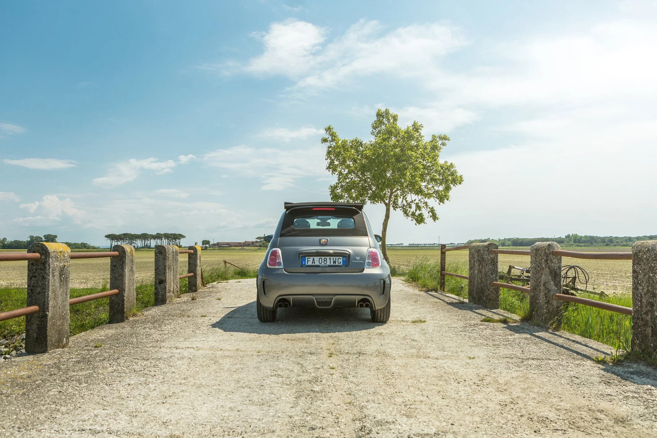 A silver car parked on a gravel driveway in front of a small tree and a wooden fenced area. The background shows a wide open field with some trees under a partly cloudy blue sky.