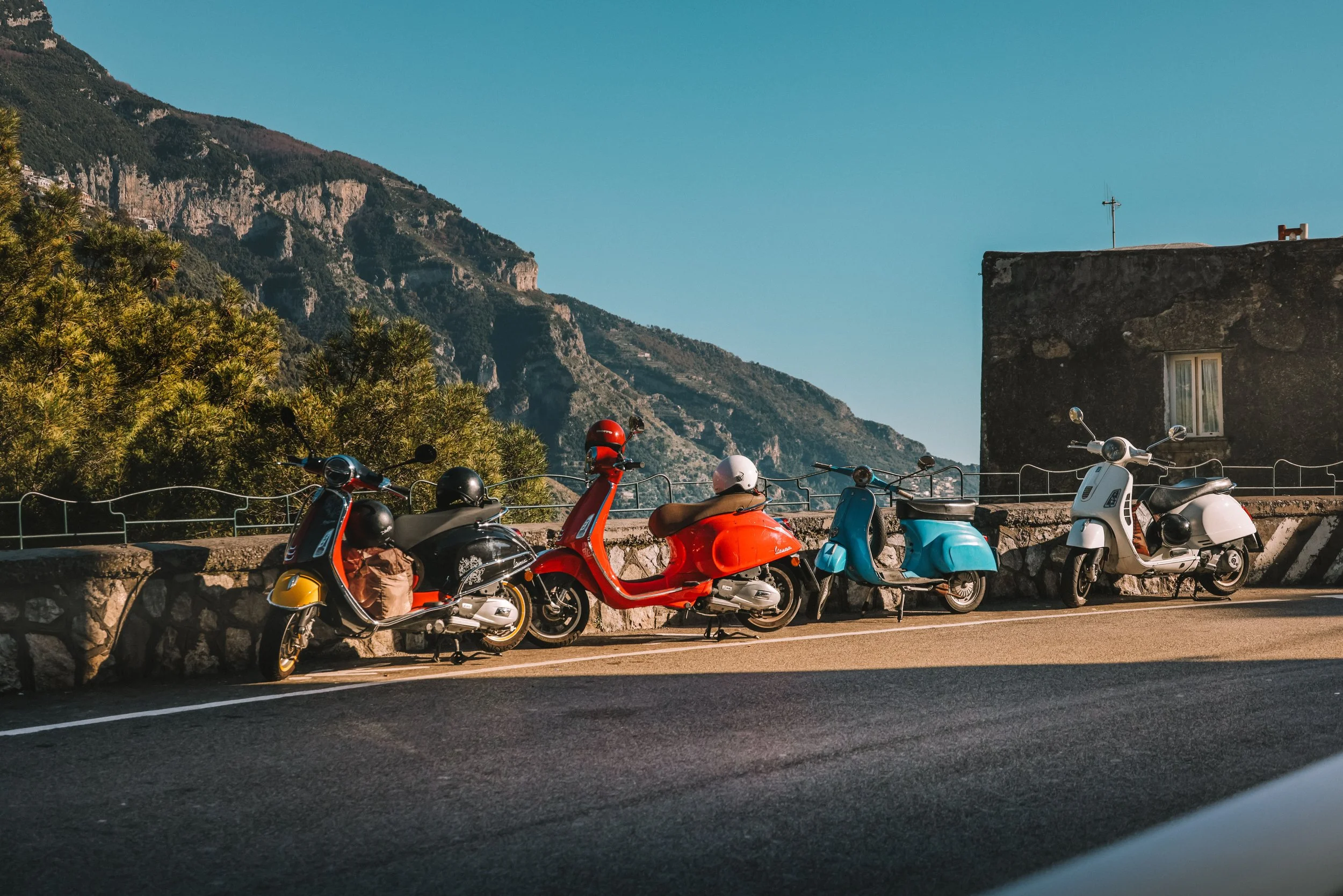 Multiple scooters and motorcycles parked along a mountain roadside with a building, trees, and mountains in the background under a clear blue sky.