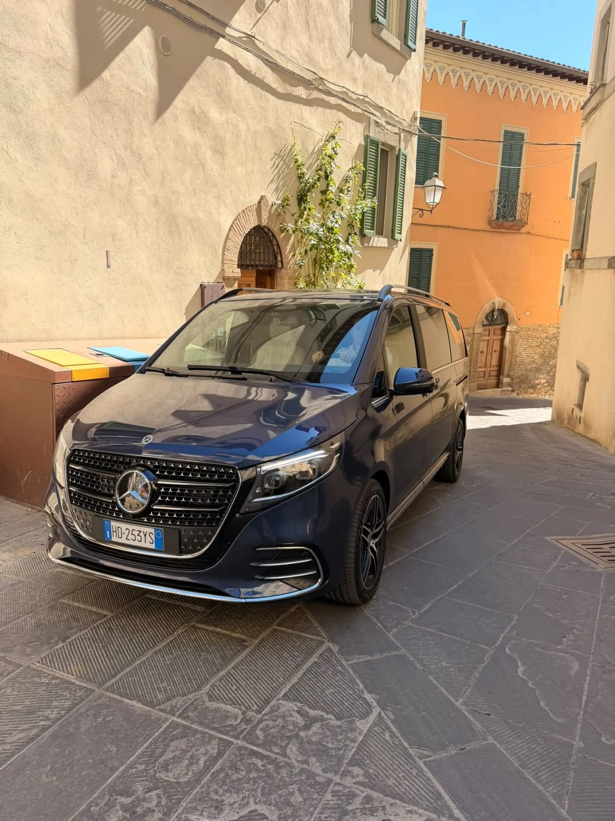 A black Mercedes-Benz SUV parked on a narrow street with colorful buildings, shutters, a lamp, and a tree in the background.