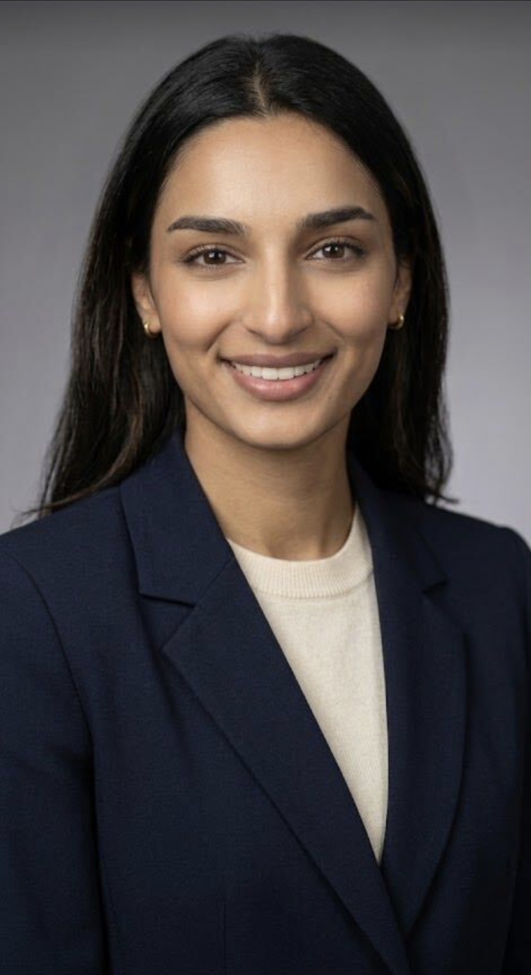 A woman with medium skin tone and dark brown hair smiling, wearing a navy blazer over a cream-colored top, posing against a gray background.