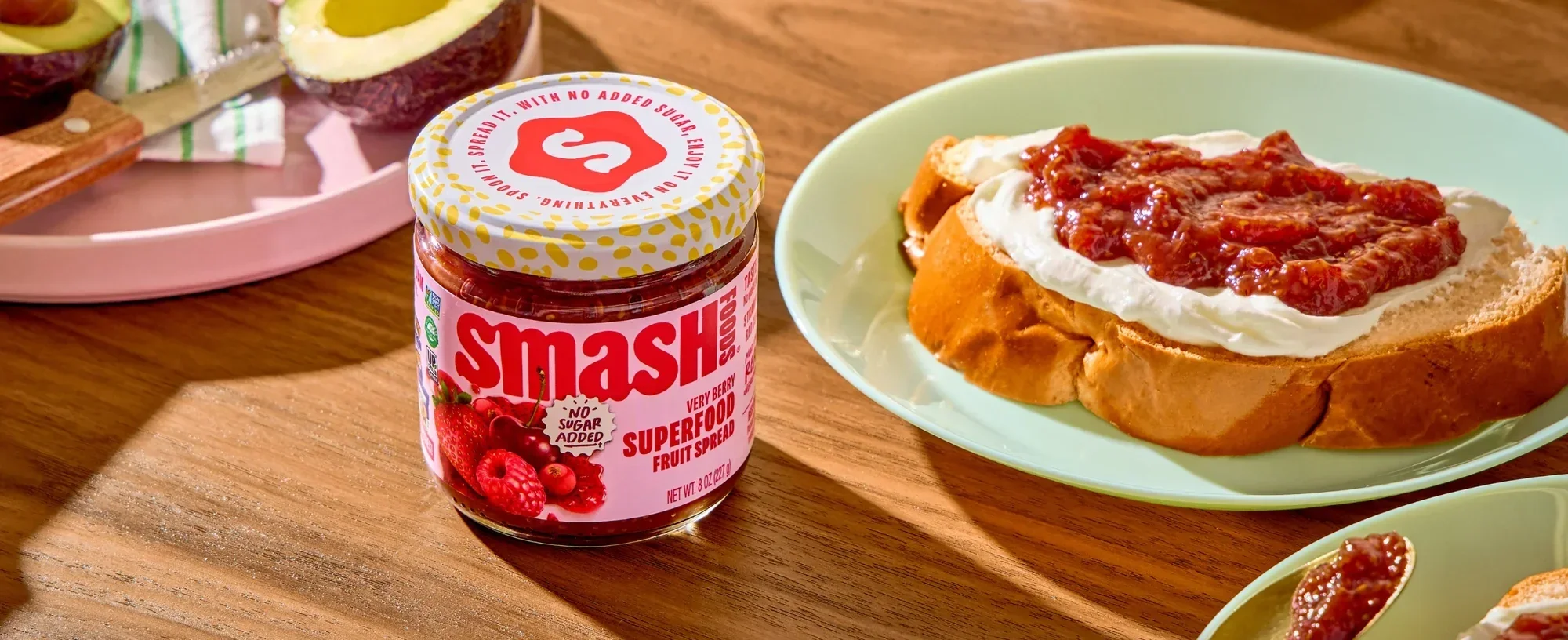 A jar of Smash superfood fruit spread and a slice of bread topped with whipped cream and fruit spread on a green plate, with part of avocados and a pink tray in the background on a wooden table.