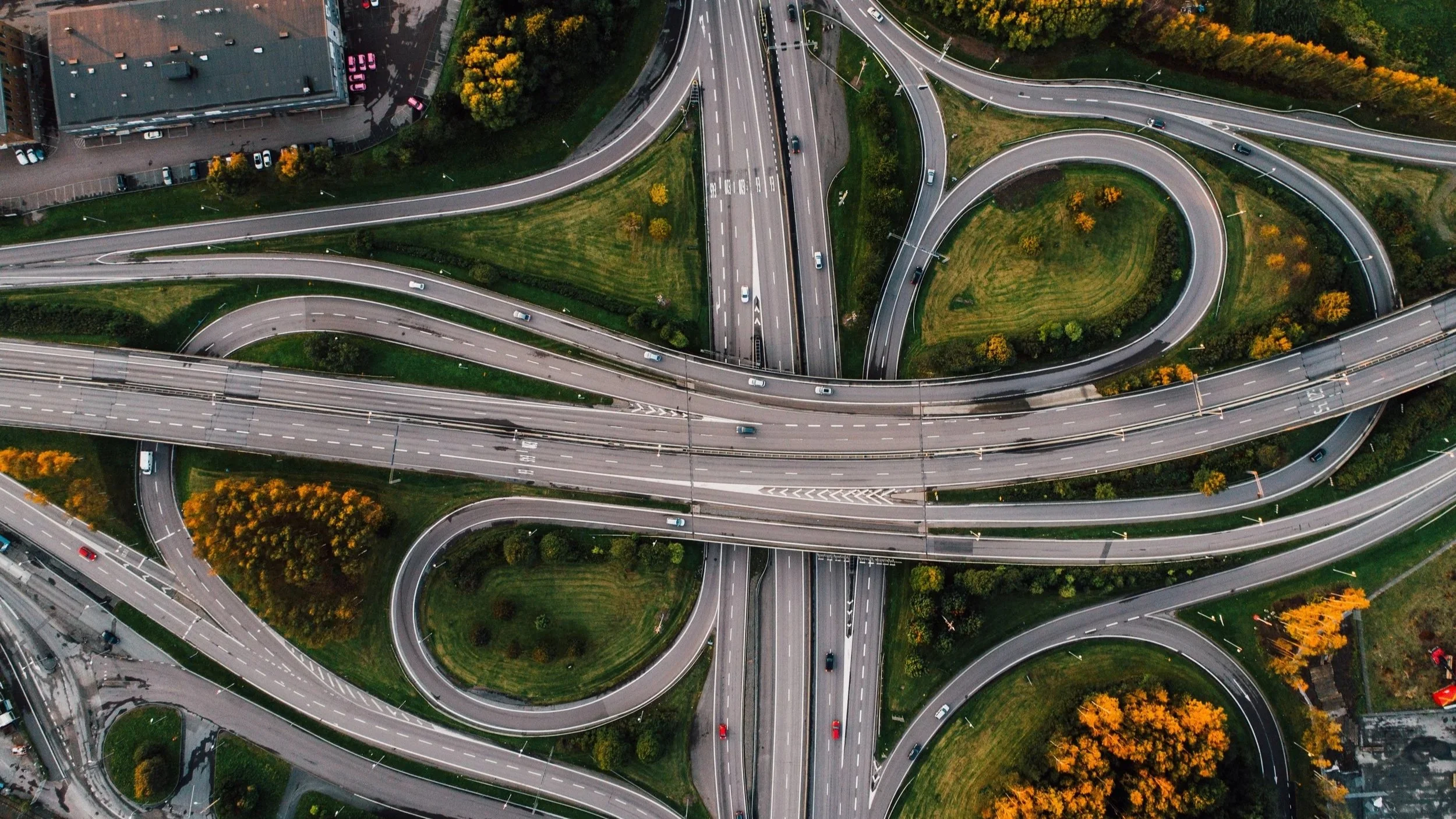 Aerial view of a complex highway interchange with multiple looping ramps, green spaces, and trees with autumn foliage.