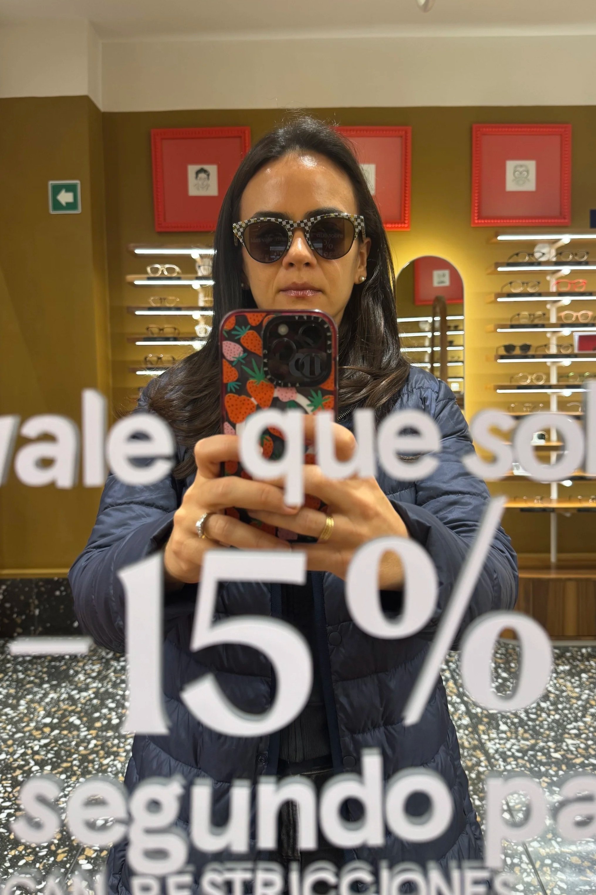 A woman taking a selfie in a store mirror, wearing sunglasses with a checkered frame and a dark jacket. Behind her are shelves with sunglasses and red framed pictures on the yellow wall.