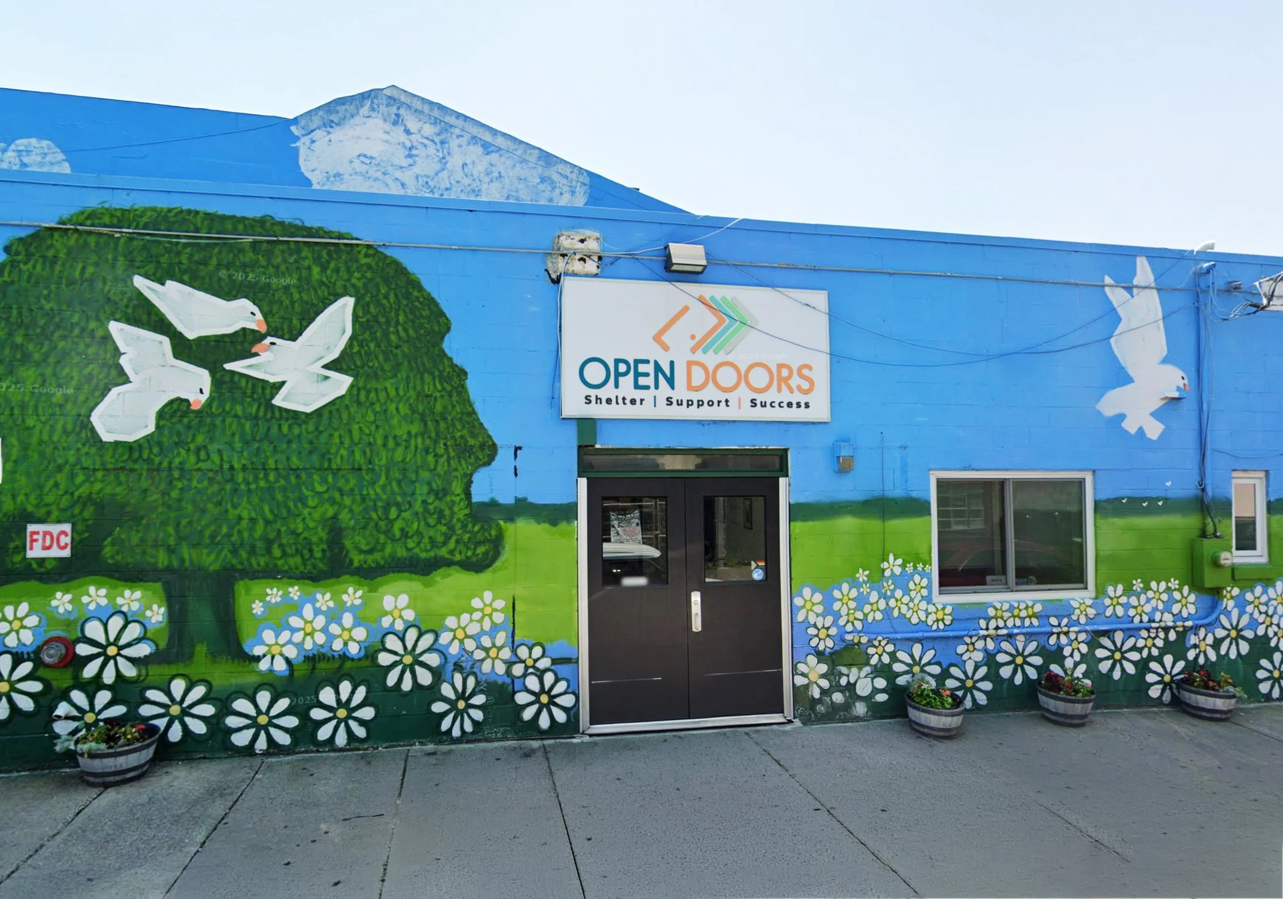Colorful mural on building wall featuring white doves, a green tree, and blue sky with mountains, with a sign that reads 'Open Doors' and the words 'Shelter | Support | Success'.