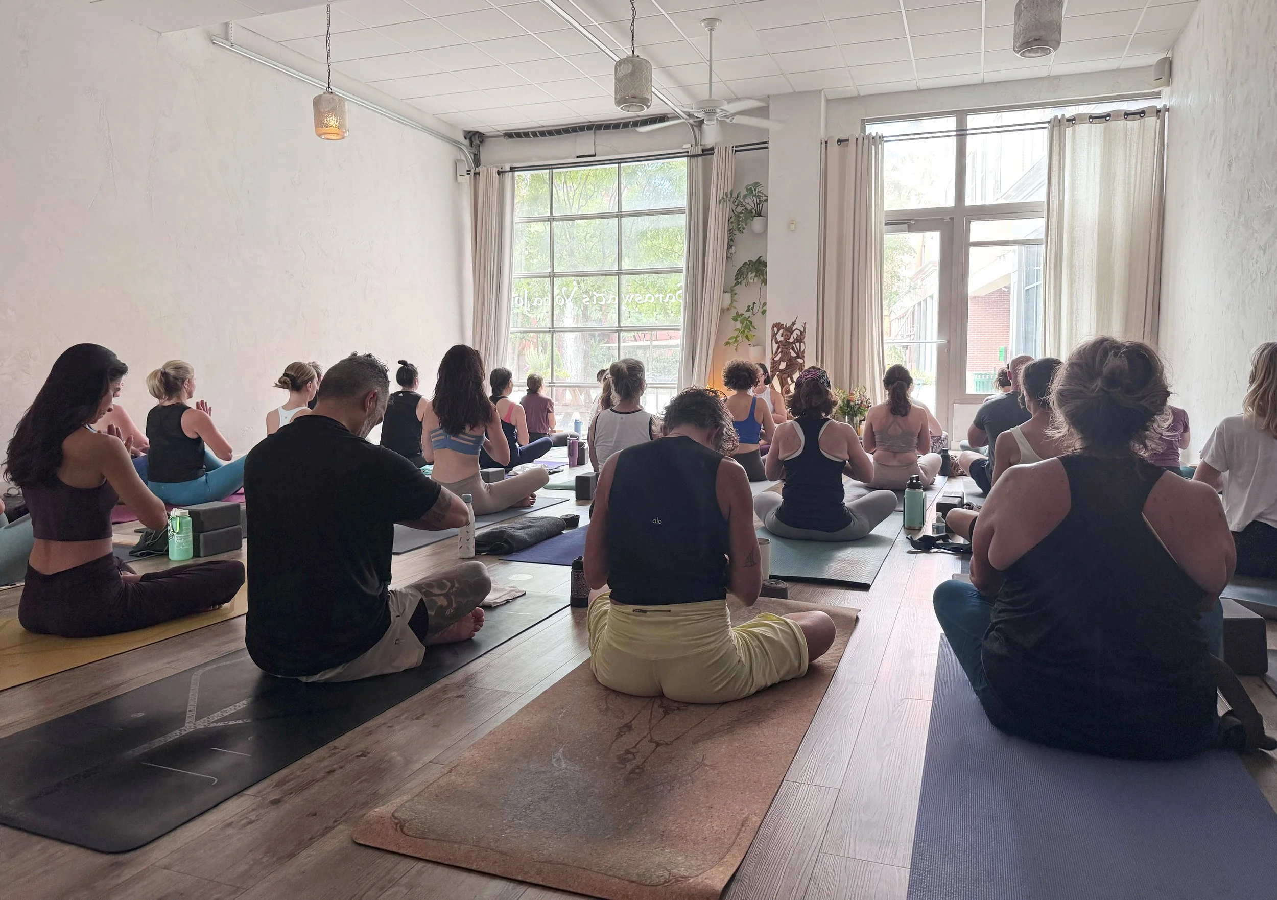Group of people practicing yoga or meditation in a bright room with large windows, curtains, and decorative plants.