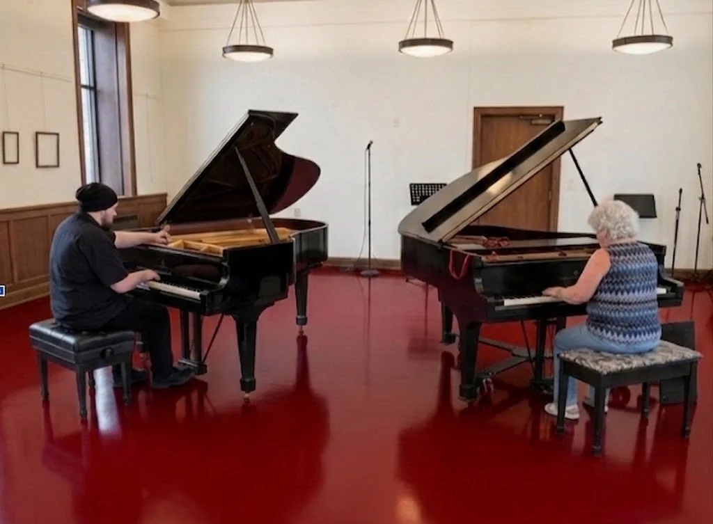 James and Annette tuning grand pianos in a room with wooden paneling and red flooring.