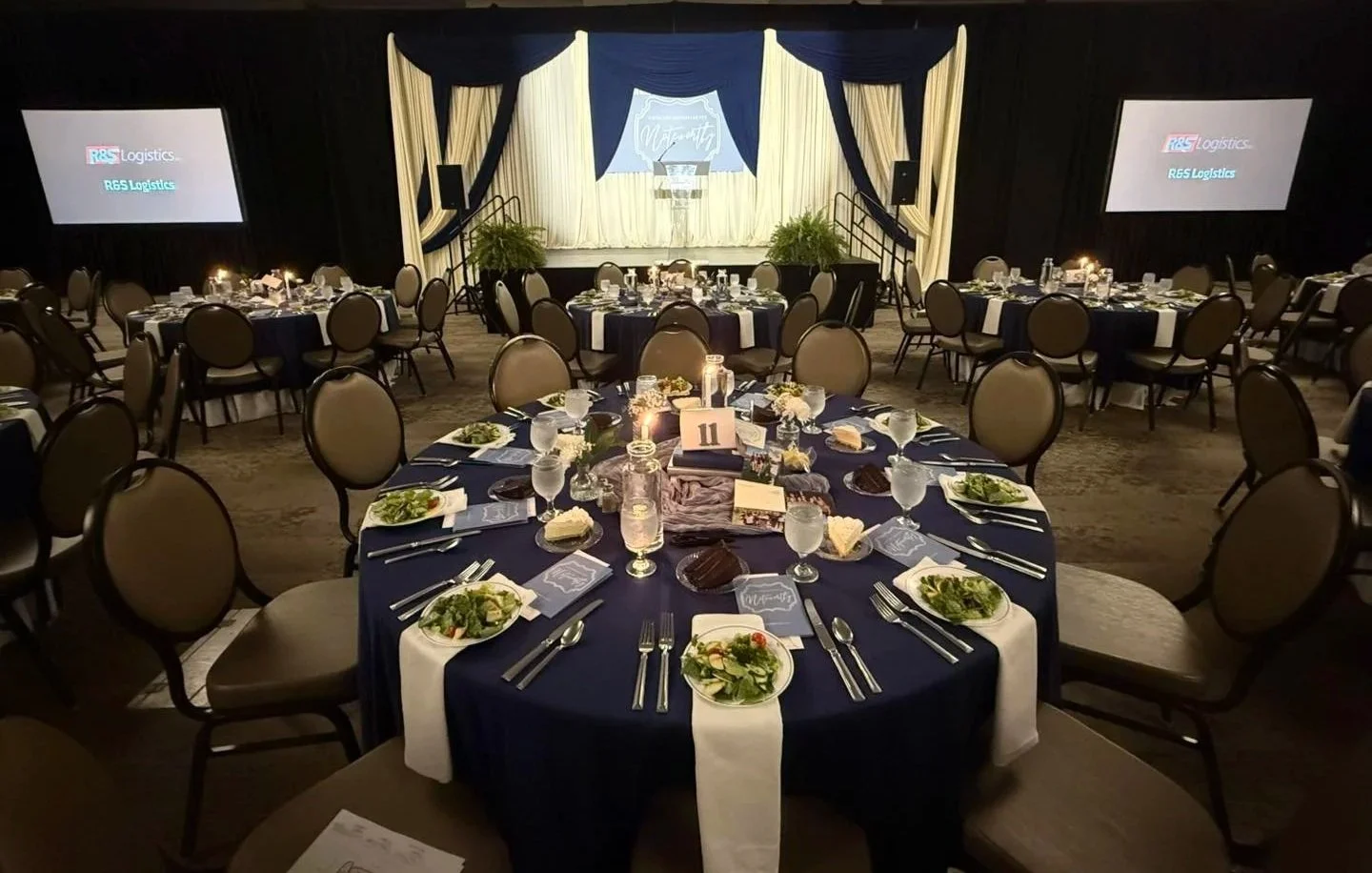 A banquet hall with round tables set for a formal event, featuring navy blue tablecloths, white napkins, salad plates, water glasses, and floral centerpieces, with a stage at the front flanked by large curtains and two projection screens on either side