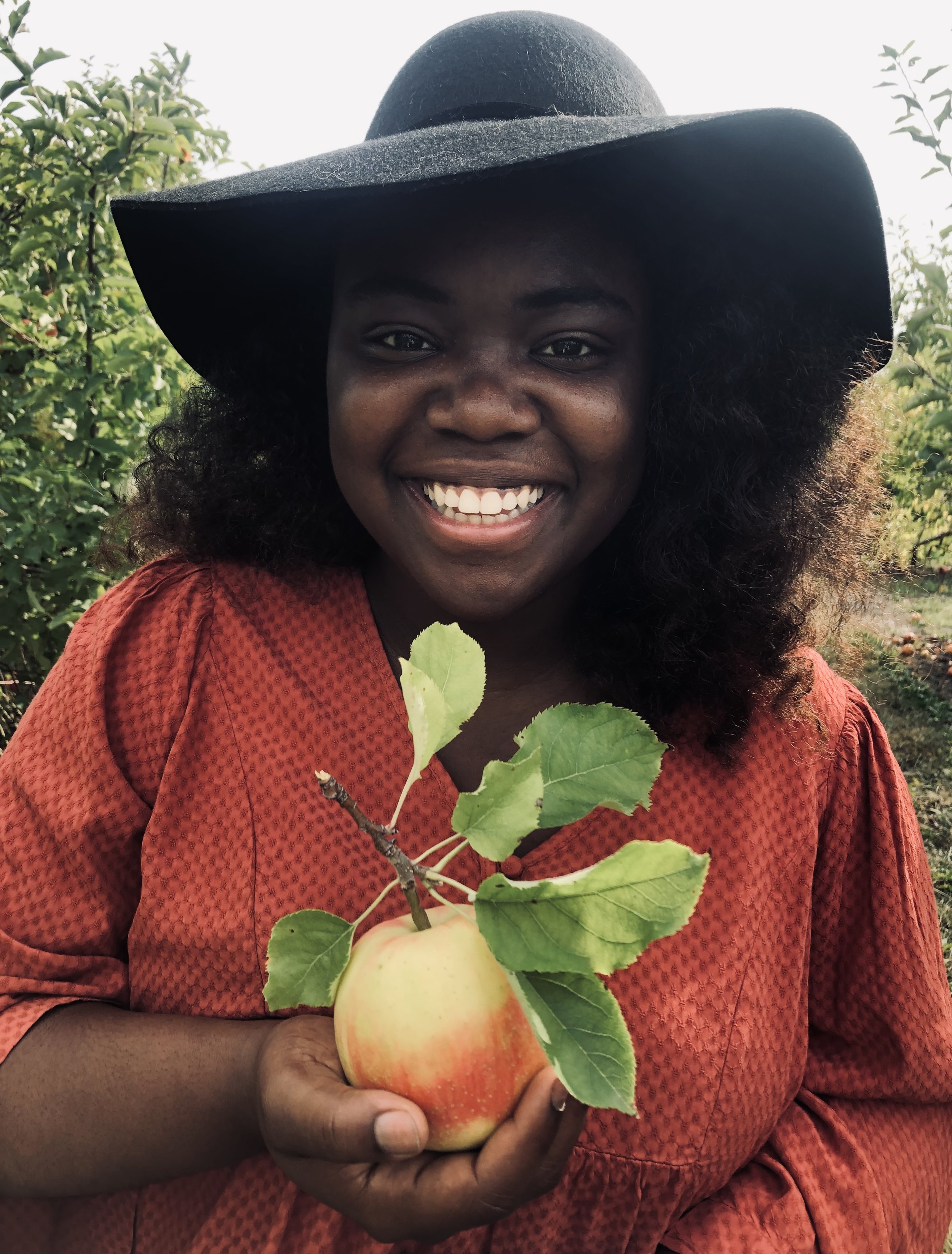 A smiling woman holding a ripe apple with green leaves, wearing a large black hat and an orange patterned top outdoors with green trees in the background.