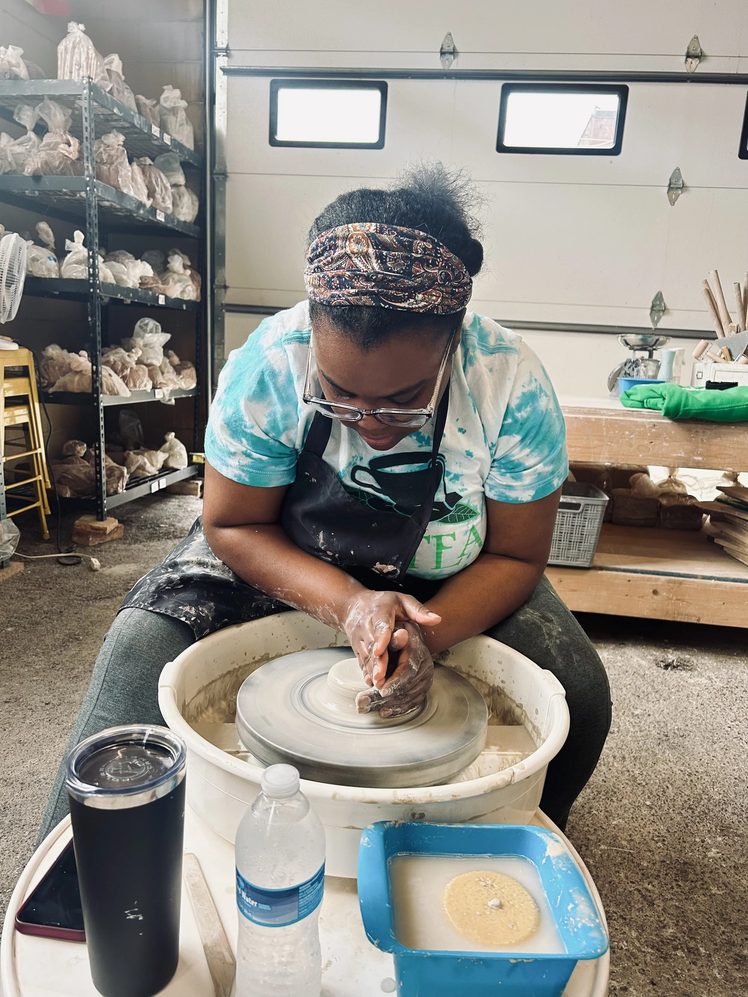 A woman shaping clay on a pottery wheel in a workshop. Shelves with bags of clay, various tools, and supplies are in the background.