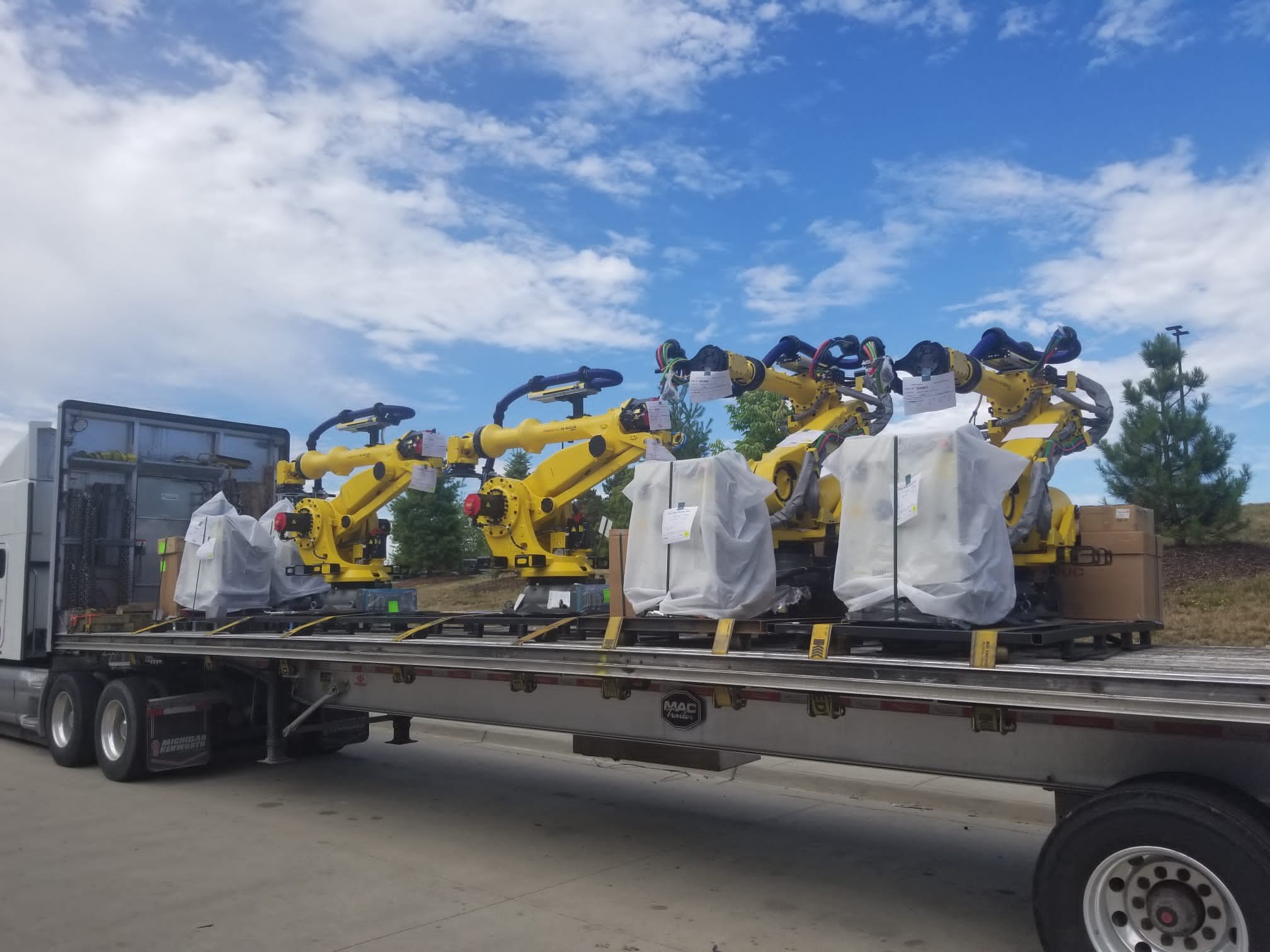 A flatbed truck transporting multiple yellow industrial robotic arms, with some wrapped in protective covering, parked outdoors under a blue sky with scattered clouds.