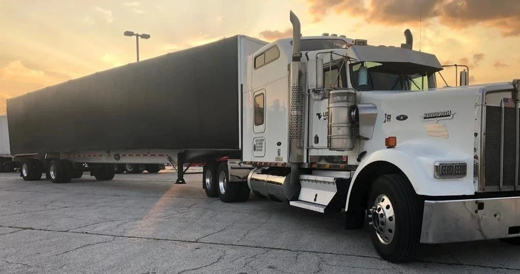 White semi-truck with a black trailer parked on a cracked asphalt lot during sunset.