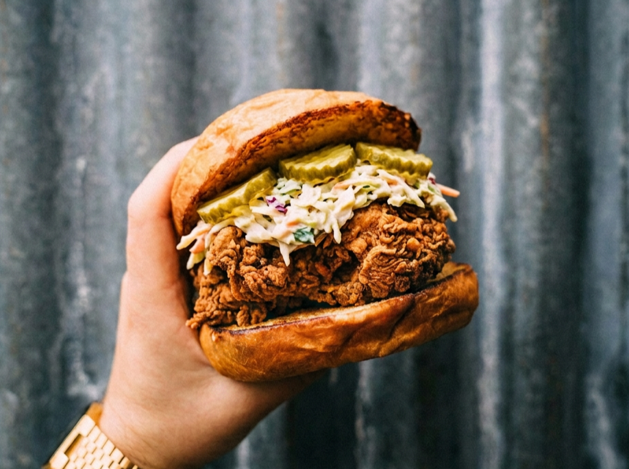 Close-up of a fried chicken sandwich with pickles, coleslaw, and a toasted bun held in a person's hand in front of a curtain background.