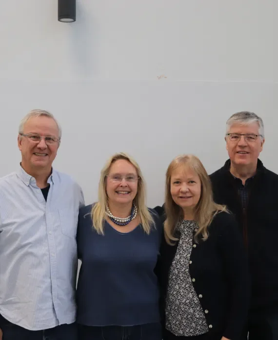 Four middle-aged adults, two men and two women, standing together indoors, smiling, in front of a plain white wall.